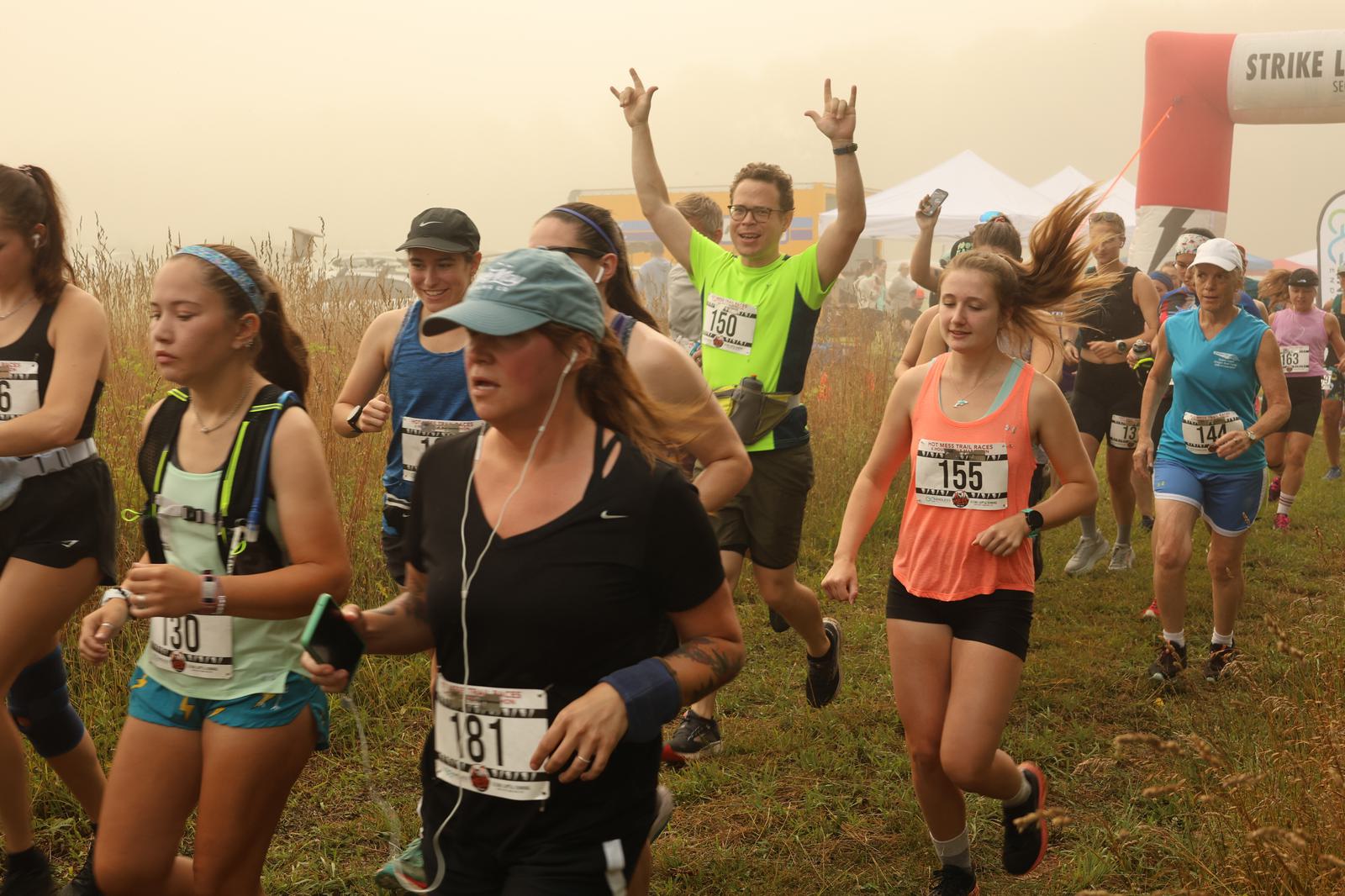 A group of runners, each wearing race bibs, jog through a grassy outdoor path in misty weather. One man in a bright green shirt raises his arms and smiles, while others focus on running. A finish arch and tents are visible in the background.
