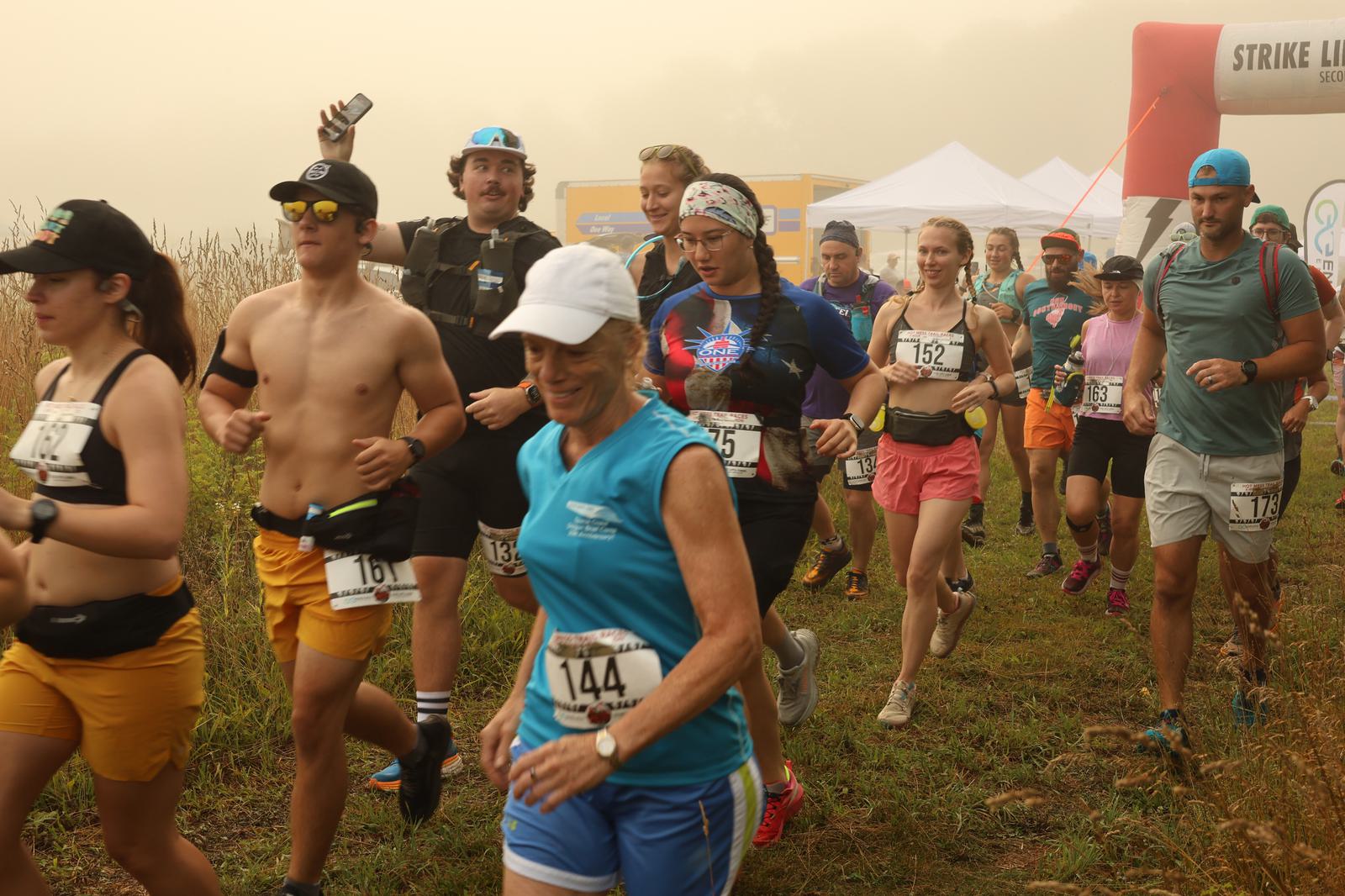 A group of runners, wearing race bibs, athletic gear, and hats, begin a race on a grassy trail near a red inflatable arch on a foggy day. Some runners smile as they move forward together.