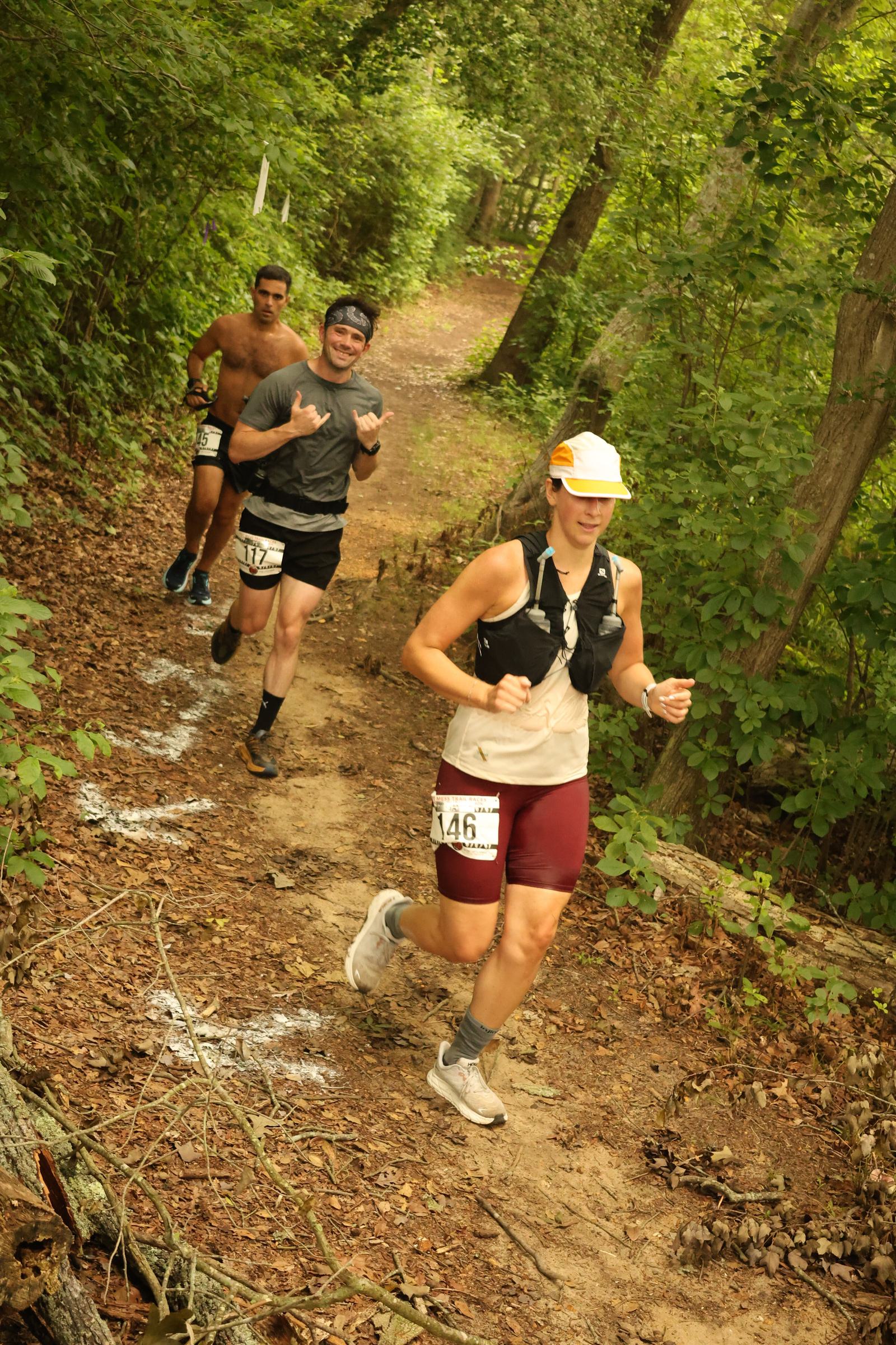 Three people run on a dirt trail through a forest. The woman in front wears a white hat and maroon shorts, and two men follow behind; one is shirtless and the other is smiling and making hand signs.