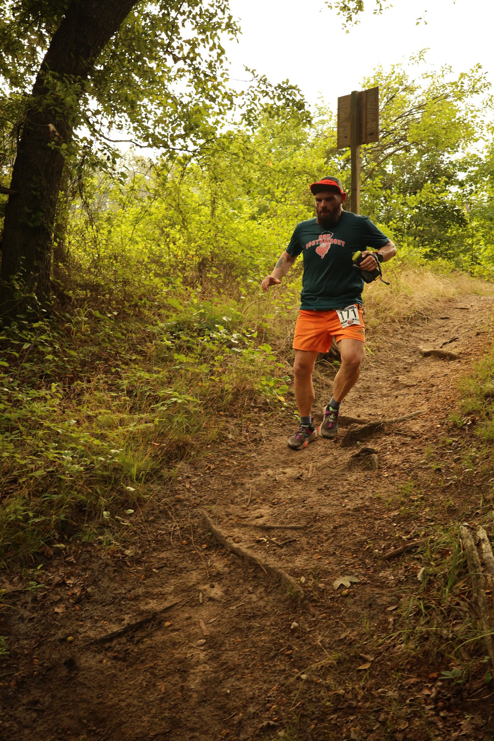 A man wearing a dark shirt, orange shorts, and a cap runs down a dirt trail in a wooded area, surrounded by trees and greenery. He is holding a bottle and appears to be participating in a race.
