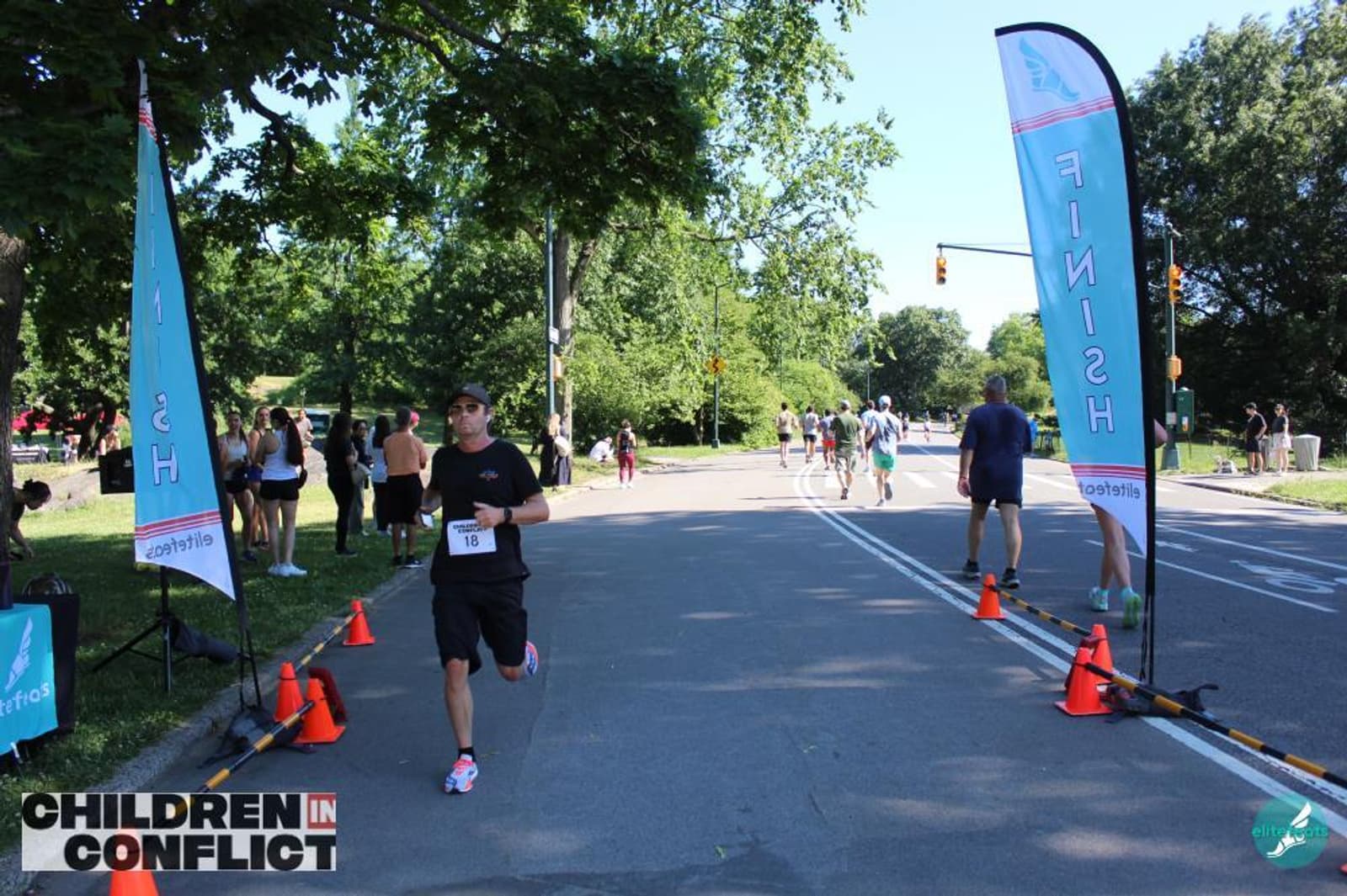 A runner approaches the finish line at an outdoor race in a park, passing between two blue flags. Spectators and other participants are visible in the background. A "Children in Conflict" logo is on the bottom left.
