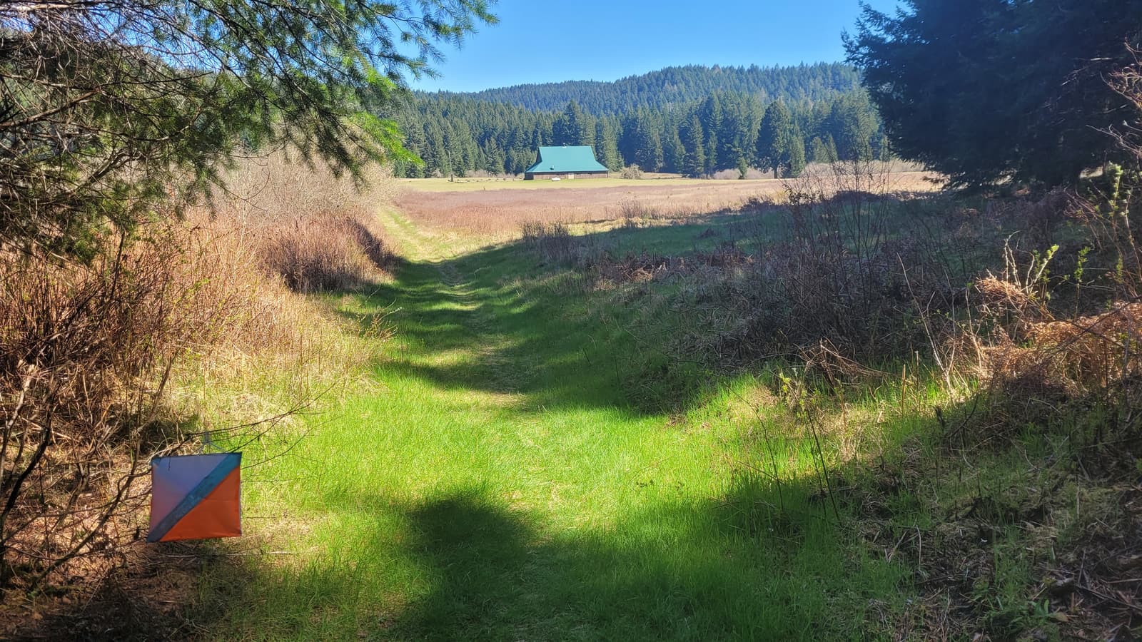 A grassy trail leads through a field toward a green-roofed building surrounded by forested hills. In the foreground, an orange and white orienteering marker sits on the left side of the path.