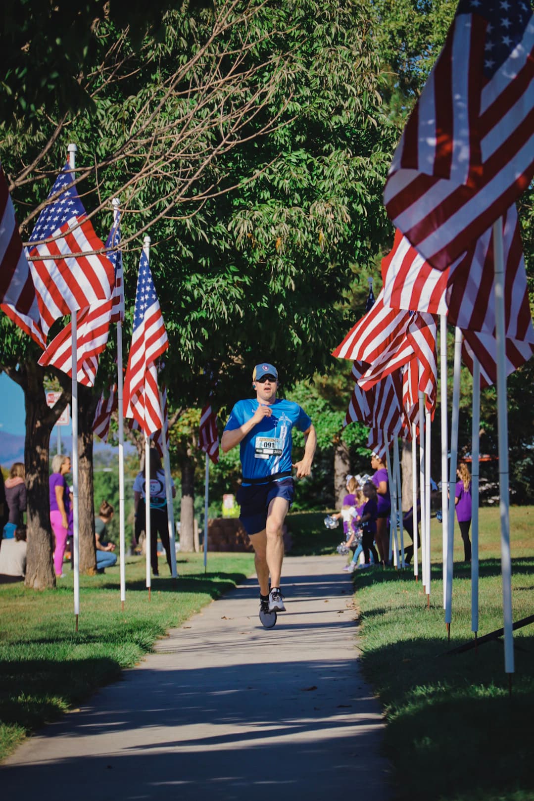 A man in blue running gear runs down a sidewalk lined with American flags on both sides in a park, with trees providing shade and people visible in the background.