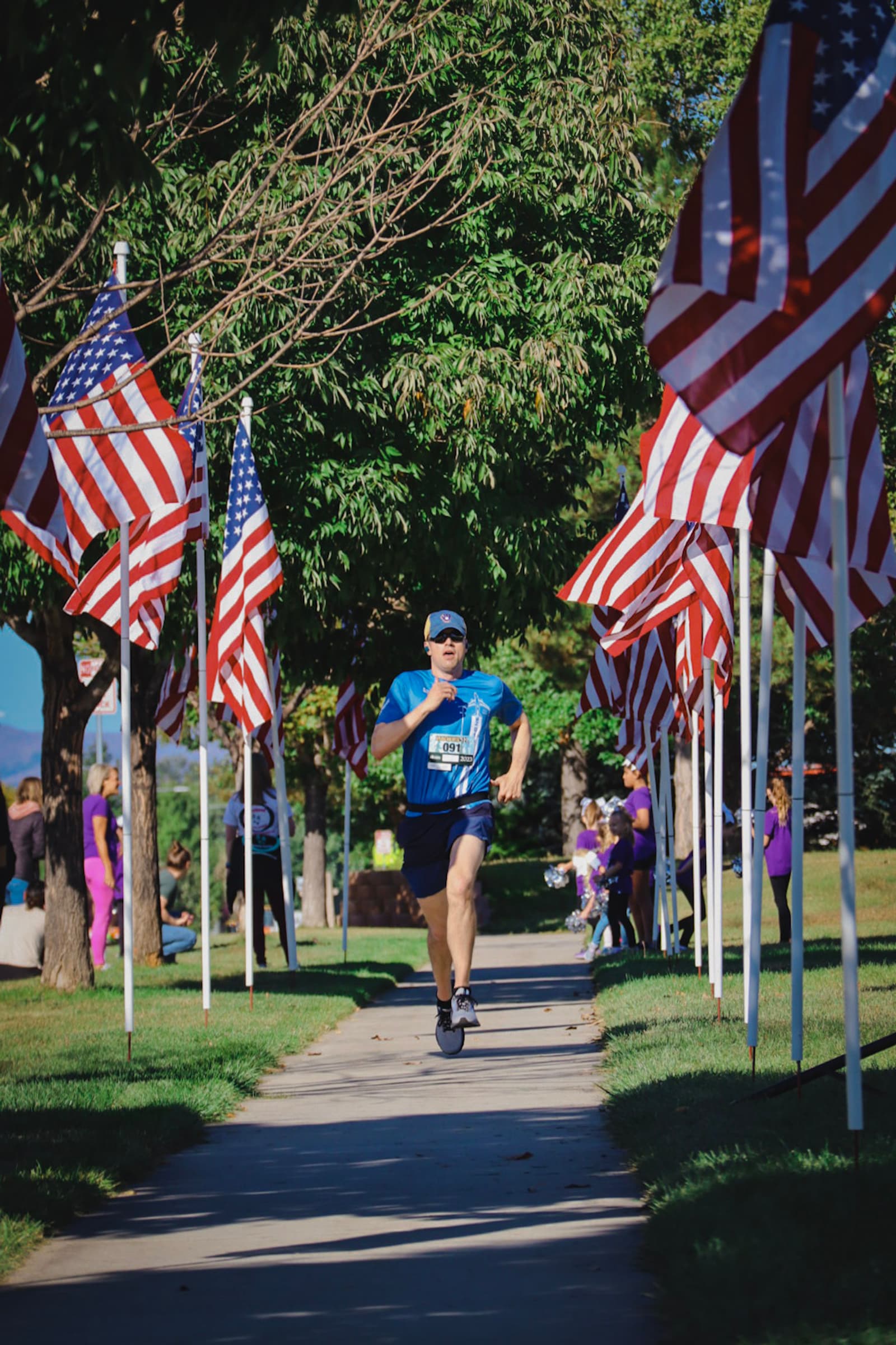 A man in blue running gear runs down a sidewalk lined with American flags on both sides in a park, with trees providing shade and people visible in the background.