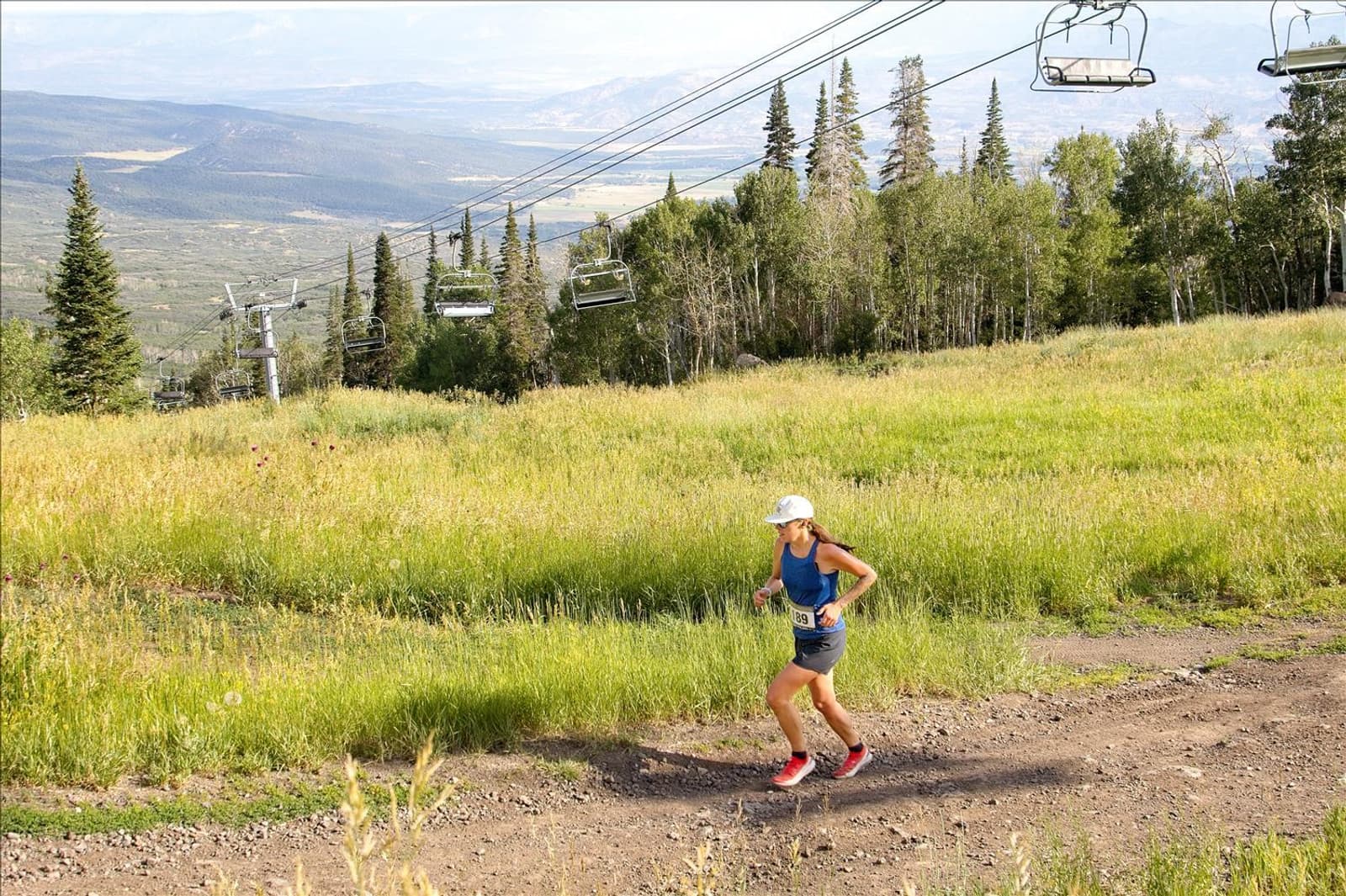 A person in running gear jogs up a dirt trail on a grassy mountain slope, with chairlifts and tall trees in the background under a partly cloudy sky.