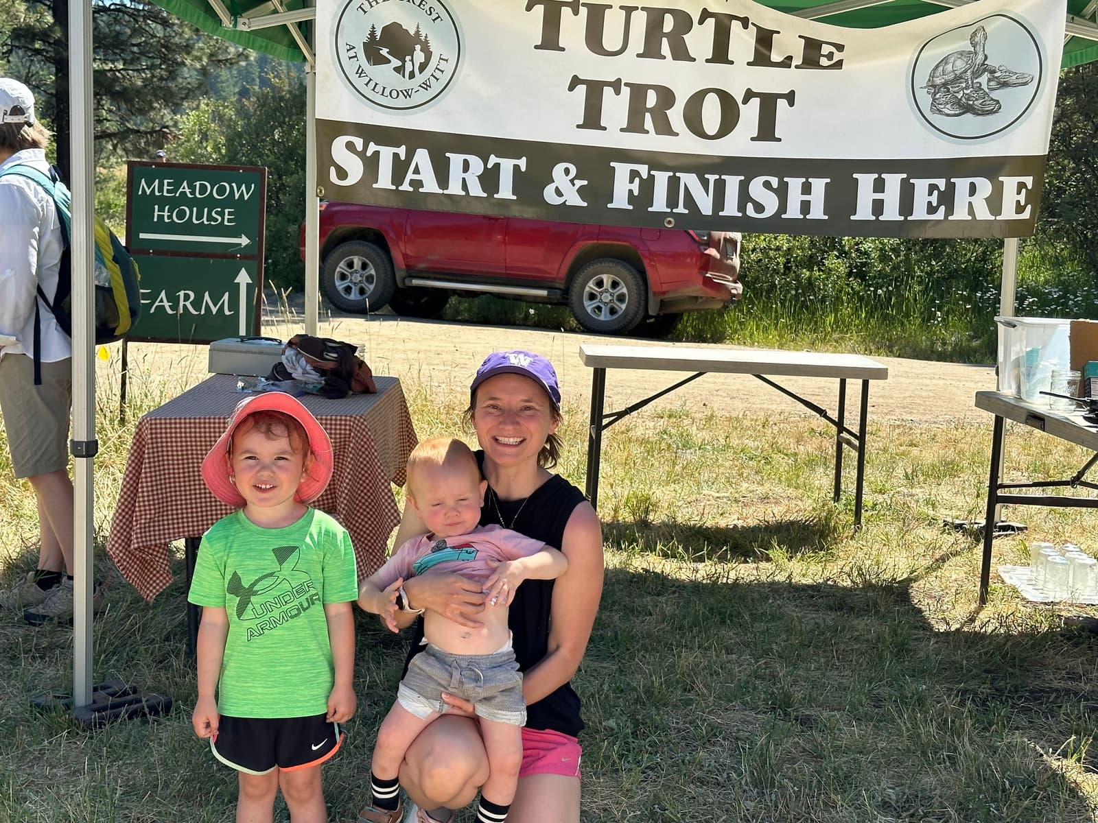 A smiling woman kneels with a baby and a young child under a "Turtle Trot Start & Finish Here" banner, with a red SUV and trees in the background on a sunny day.