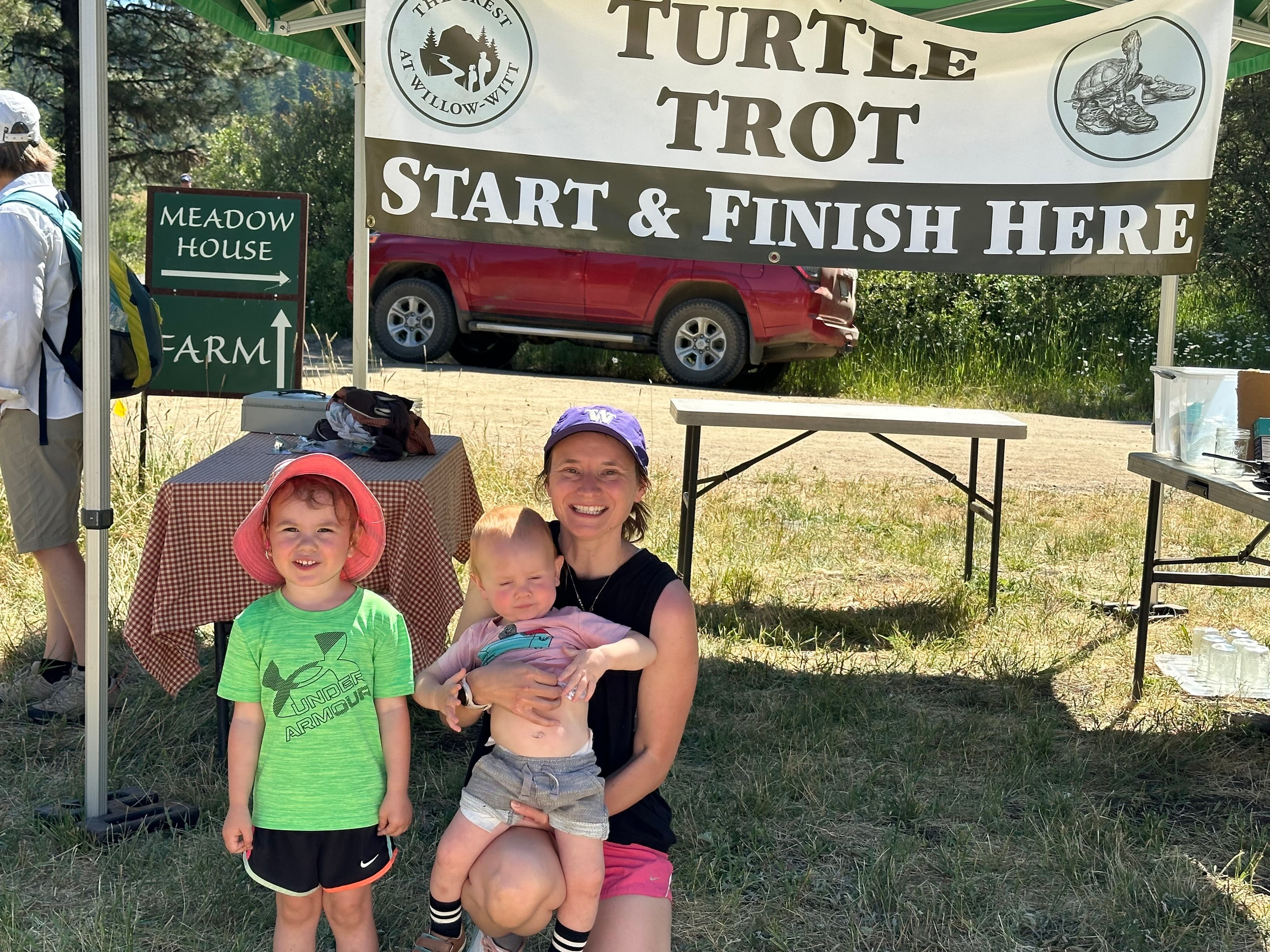 A smiling woman kneels with a baby and a young child under a "Turtle Trot Start & Finish Here" banner, with a red SUV and trees in the background on a sunny day.