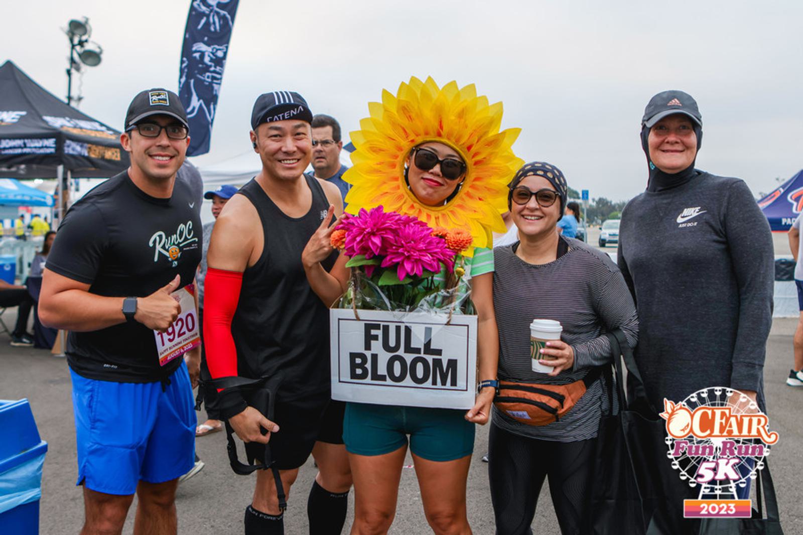Five people at a 5K fun run, one dressed as a sunflower holding a "Full Bloom" sign with flowers. All are smiling, wearing athletic gear, with event tents and the OC Fair Fun Run 5K 2023 logo in the background.