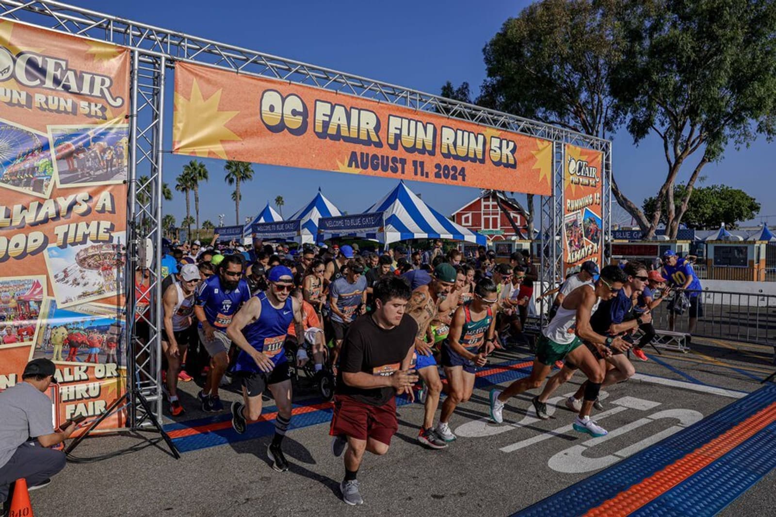 Runners of all ages start the OC Fair Fun Run 5K, passing under a large banner with the event name and date, surrounded by fair tents and palm trees on a sunny day.