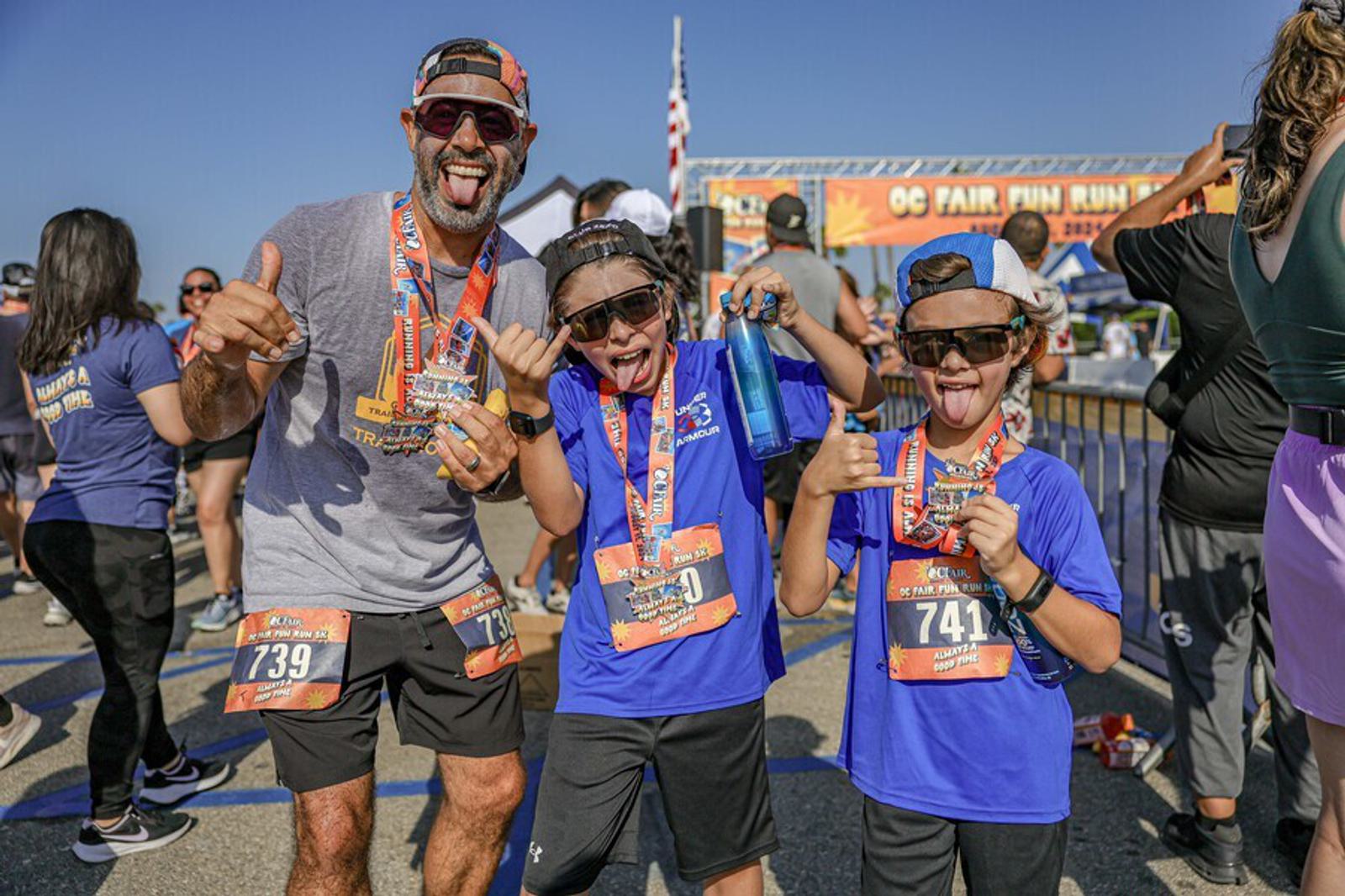 A man and two children wearing race bibs and medals pose smiling and giving thumbs-up gestures after finishing a fun run event, with other participants and an event banner in the background.
