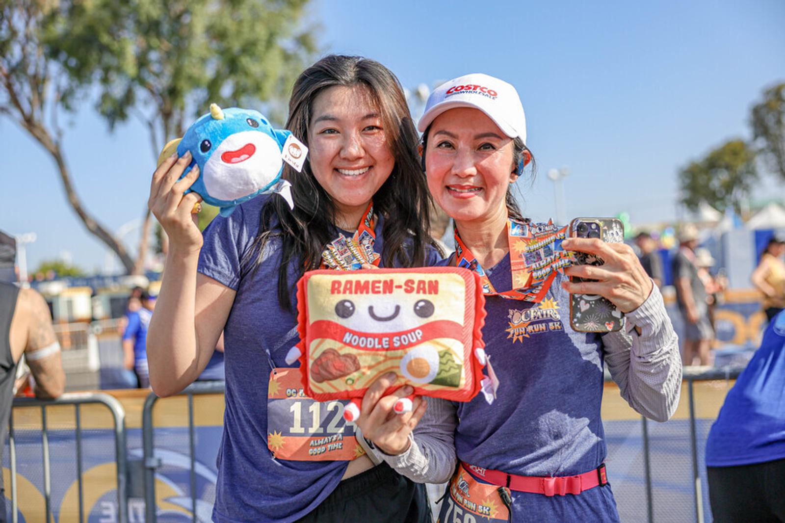 Two women smiling and holding up plush toys and medals after a race event. They wear race bibs and purple shirts, standing outdoors with other participants and trees visible in the background.