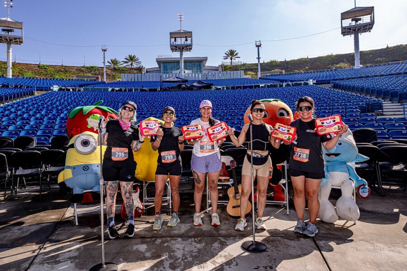 Six people in athletic wear stand on an outdoor stage holding prize boxes, surrounded by large cartoon character mascots, with empty blue stadium seats and a sunny sky in the background.