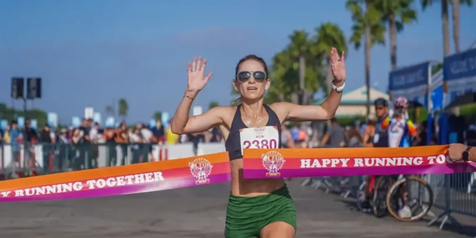 A woman wearing sunglasses and a race bib number 2380 raises her arms in victory as she crosses the finish line of a race, with a crowd and palm trees in the background.