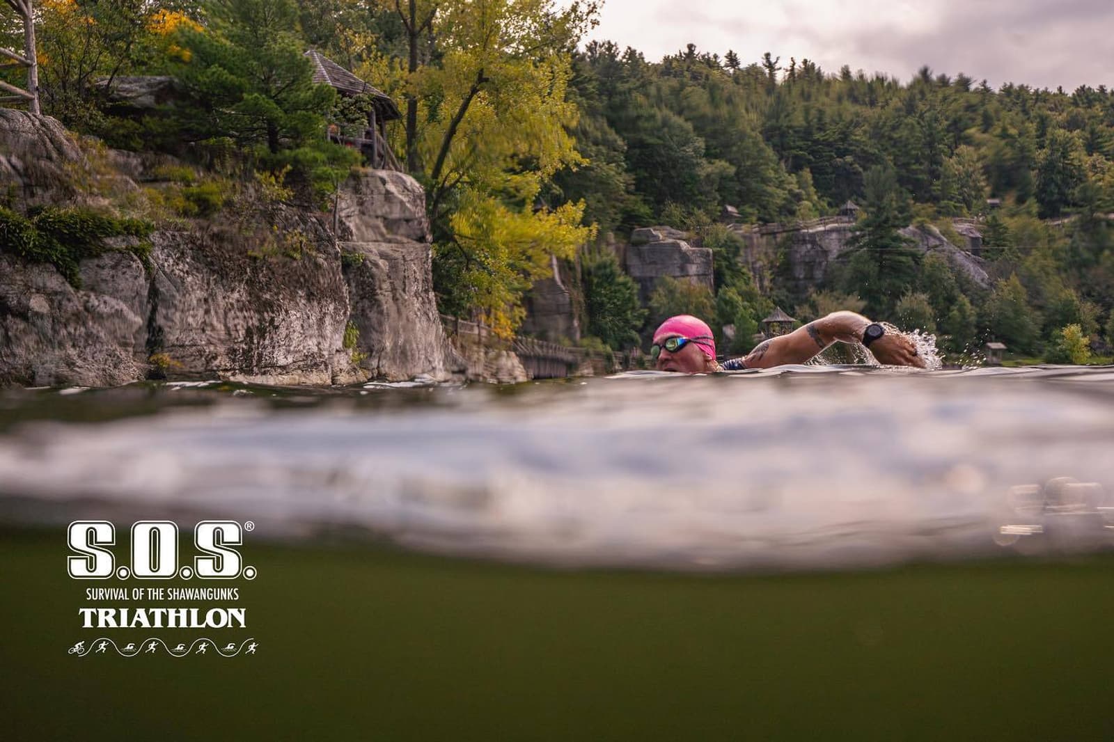 A swimmer wearing goggles and a pink swim cap moves through a lake beneath tree-lined cliffs during a triathlon event. The S.O.S. Triathlon logo is visible in the lower left corner of the image.