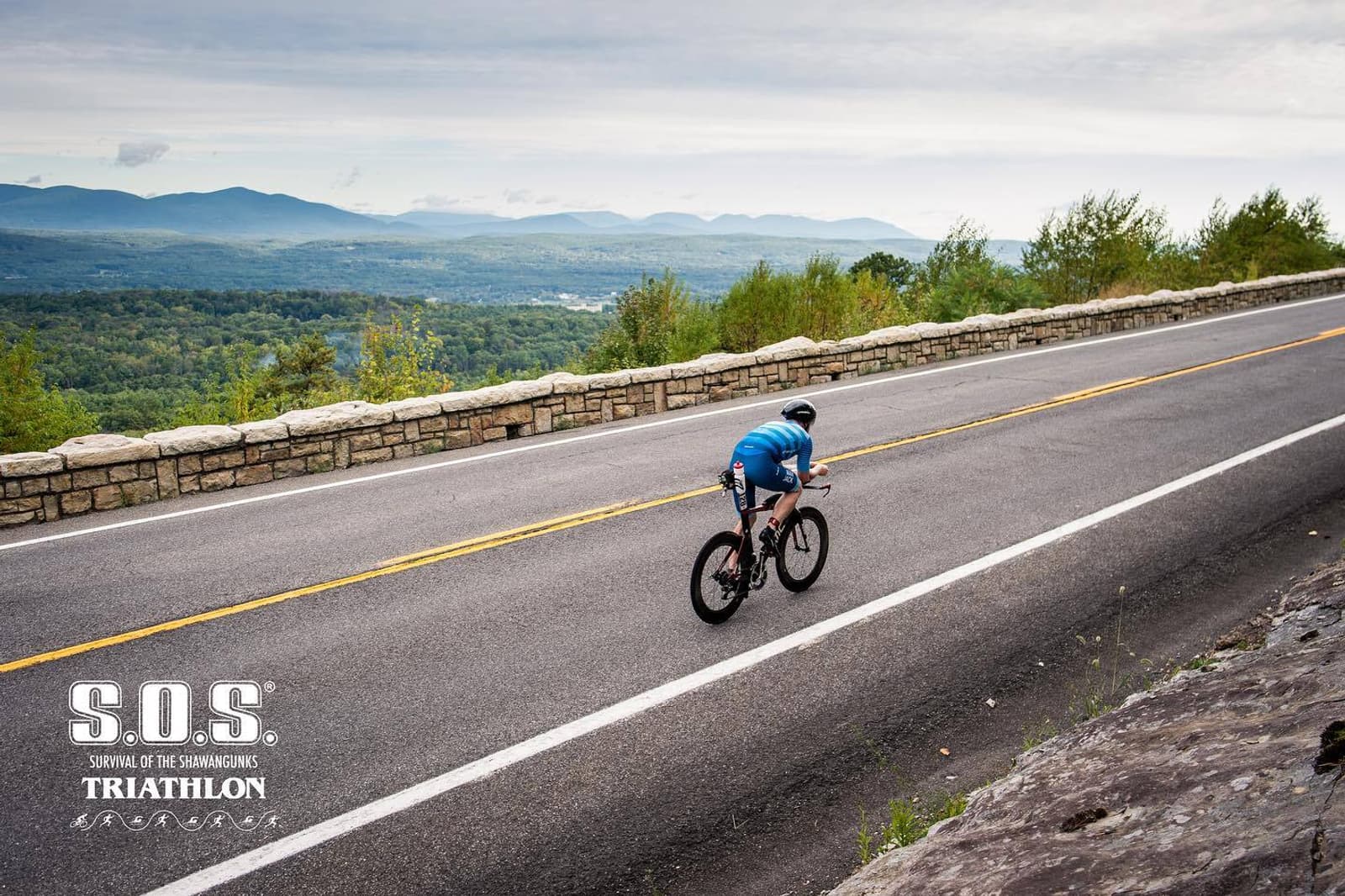 A cyclist in blue rides uphill on a scenic, empty road bordered by stone walls, with lush green hills and mountains in the background. The image includes the "S.O.S. Triathlon" logo in the lower left corner.