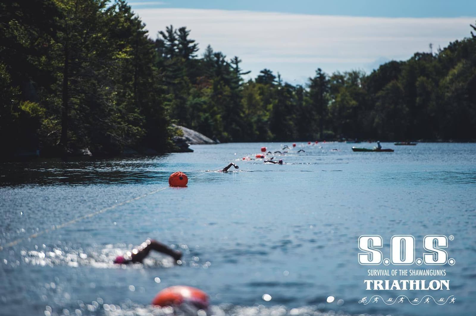 Athletes swim in open water marked by buoys, surrounded by trees under a partly cloudy sky. The S.O.S. Survival of the Shawangunks Triathlon logo is visible in the lower right corner.