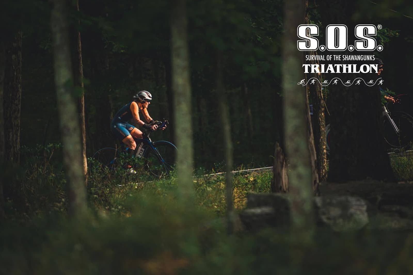 A cyclist in racing gear rides through a forested trail during the S.O.S. (Survival of the Shawangunks) Triathlon. The event logo appears in the upper right corner. Sunlight filters softly through the trees.