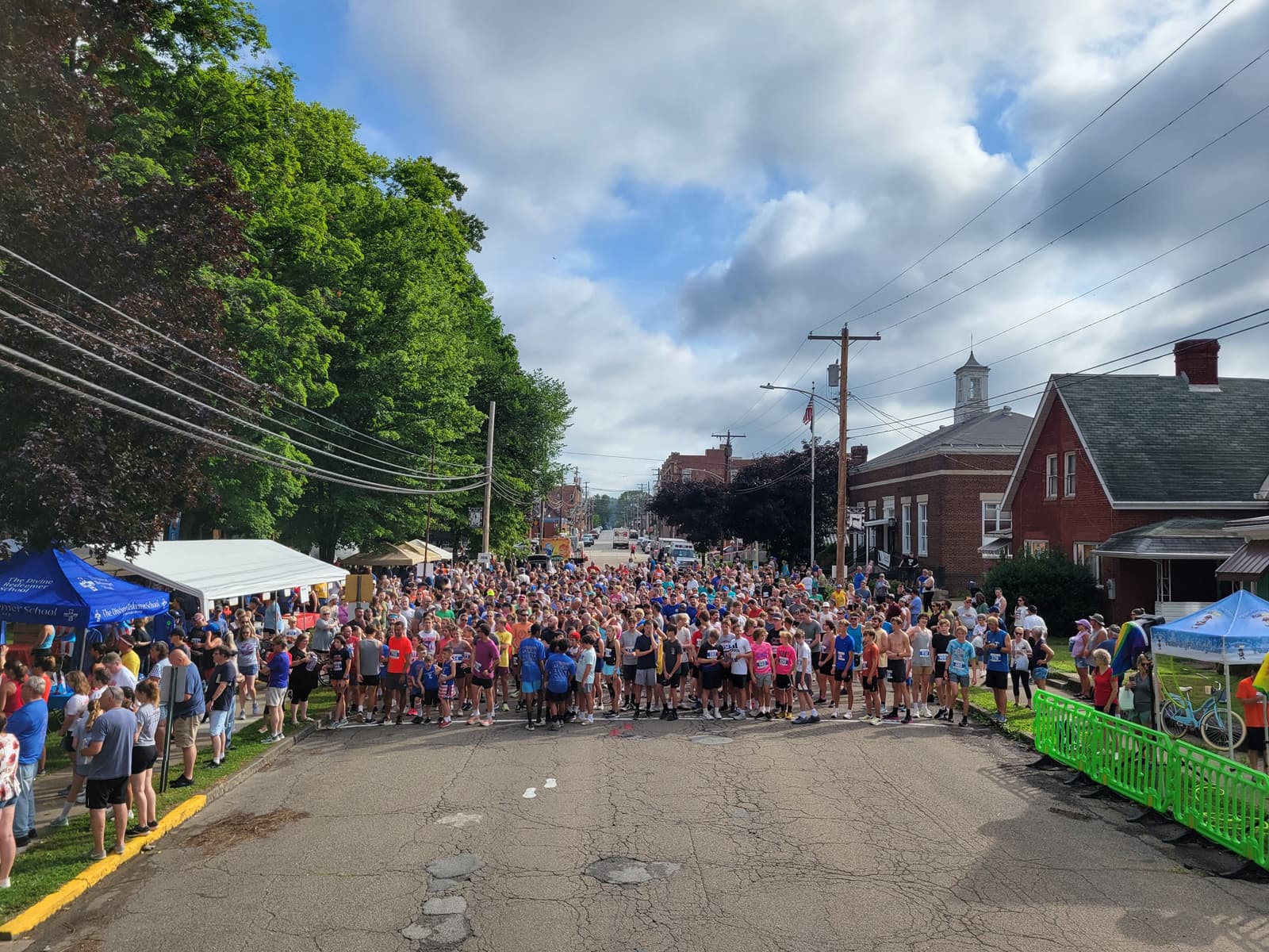 A large crowd of runners gathers at the starting line of a road race on a sunny day, with spectators and tents lining both sides of the street in a small town setting.