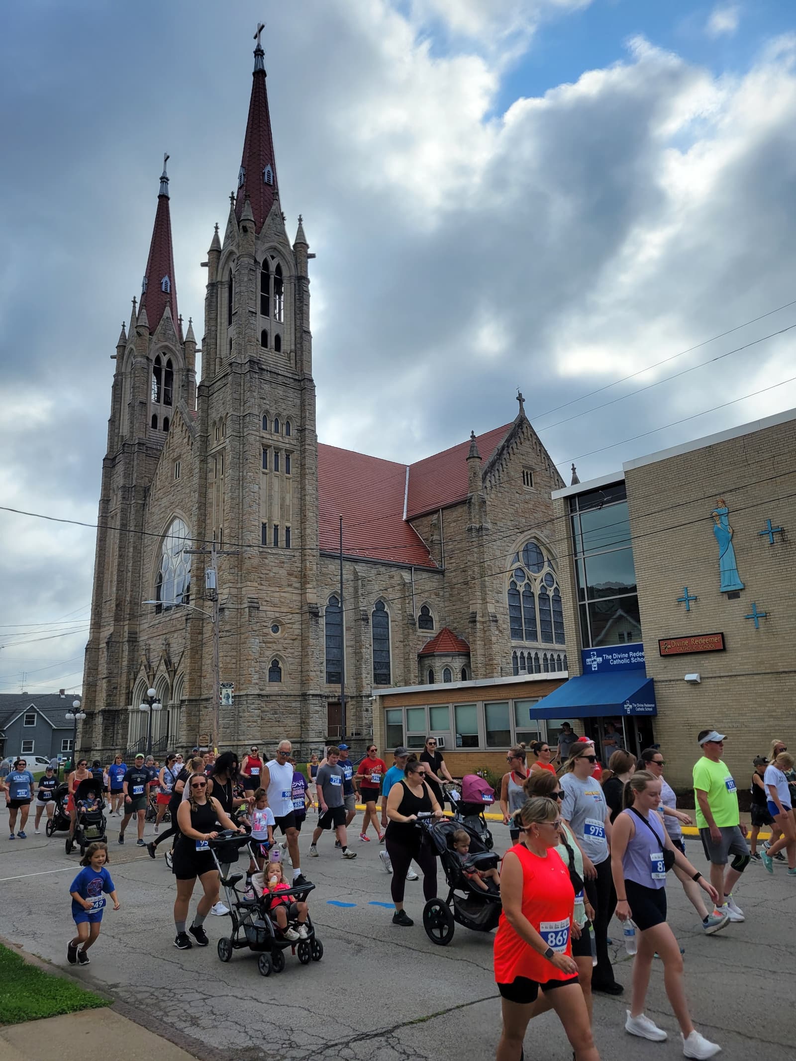 A group of people, some pushing strollers, participate in a community race or walk past a large stone church with twin spires under a partly cloudy sky.