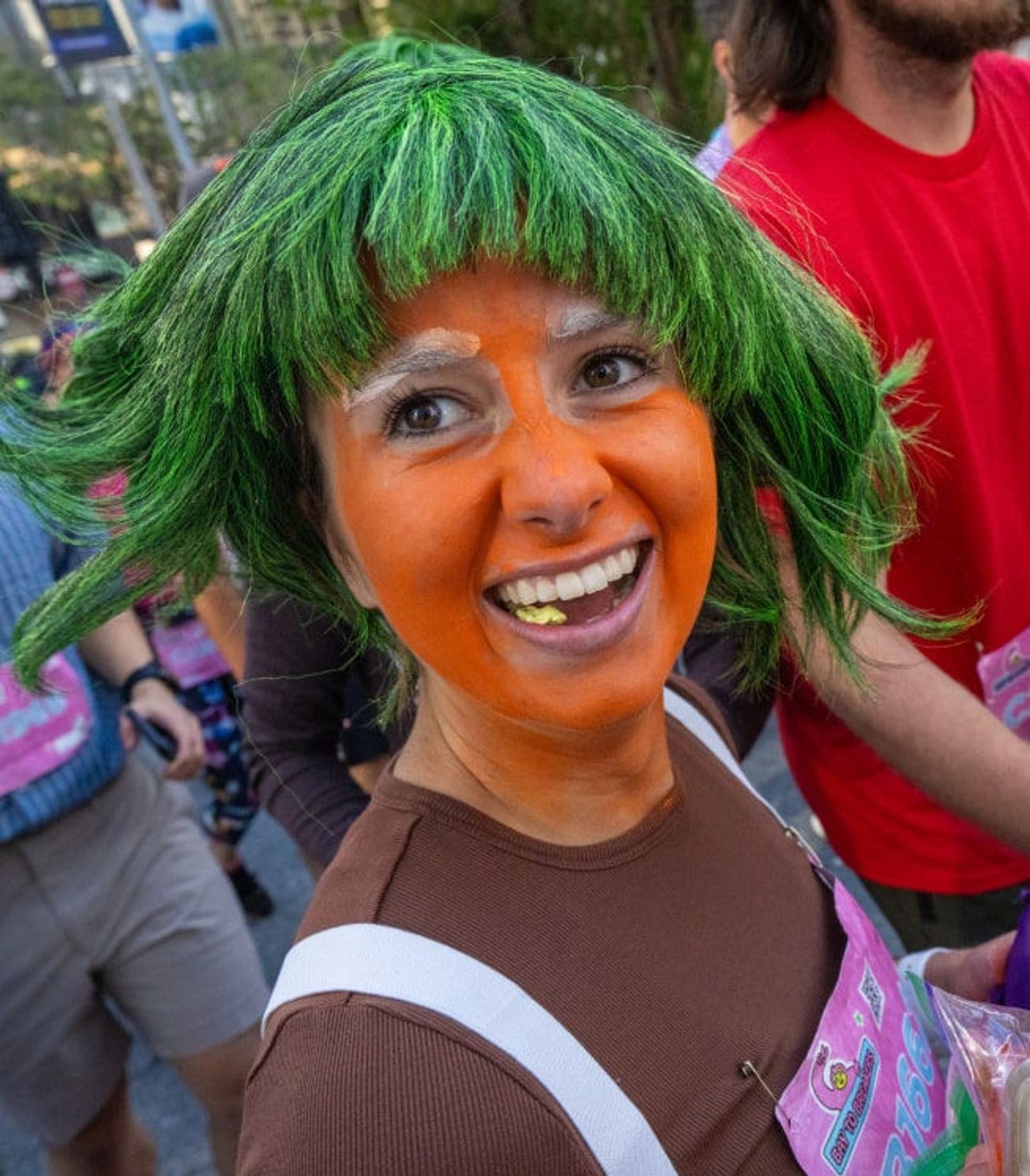 A smiling person with bright green hair and orange face paint, dressed in a brown outfit, stands among a crowd outdoors, appearing to participate in a festive or themed event.