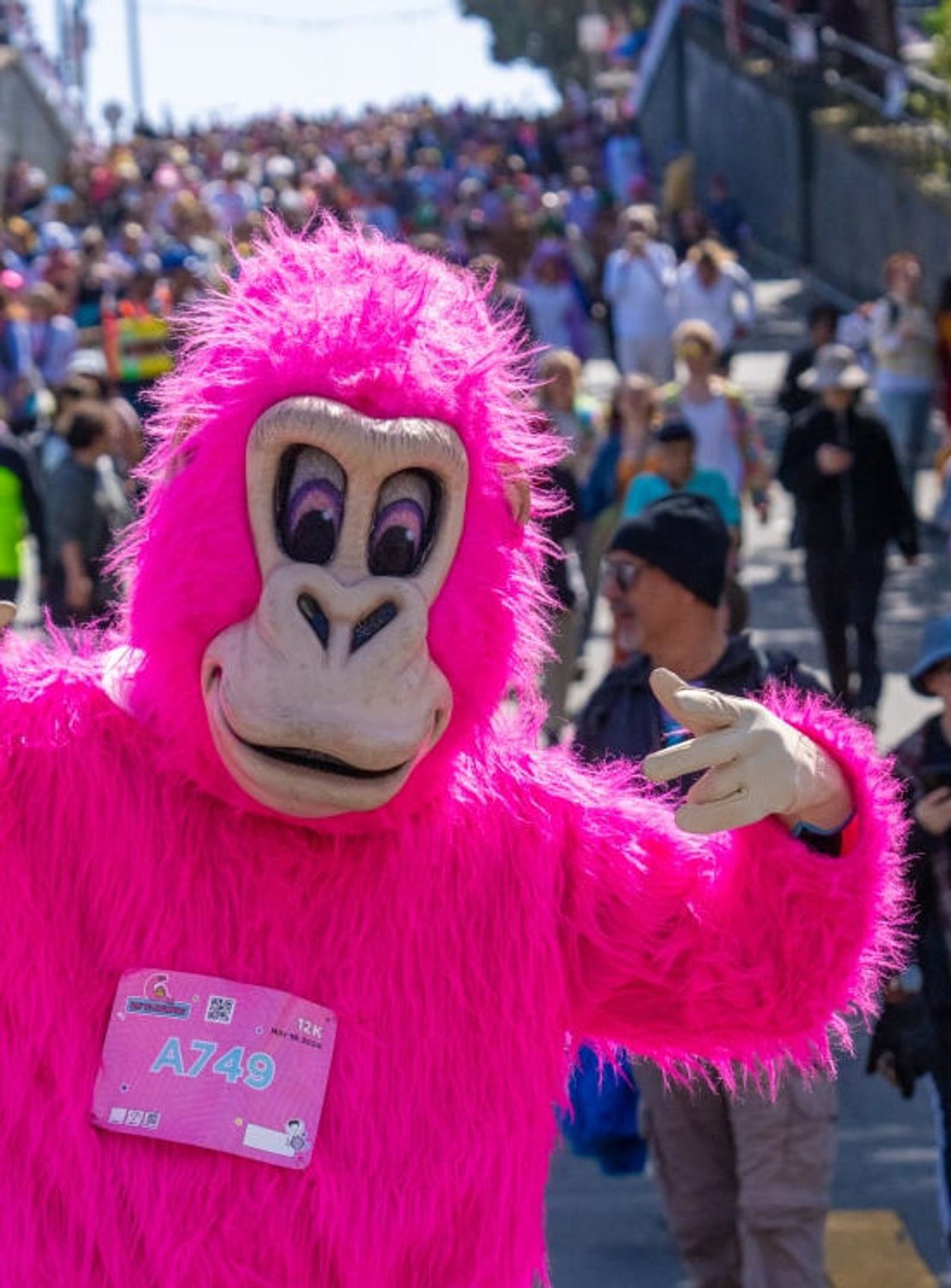A person wearing a bright pink gorilla costume poses and points towards the camera at an outdoor event, with a large crowd of people in the background.