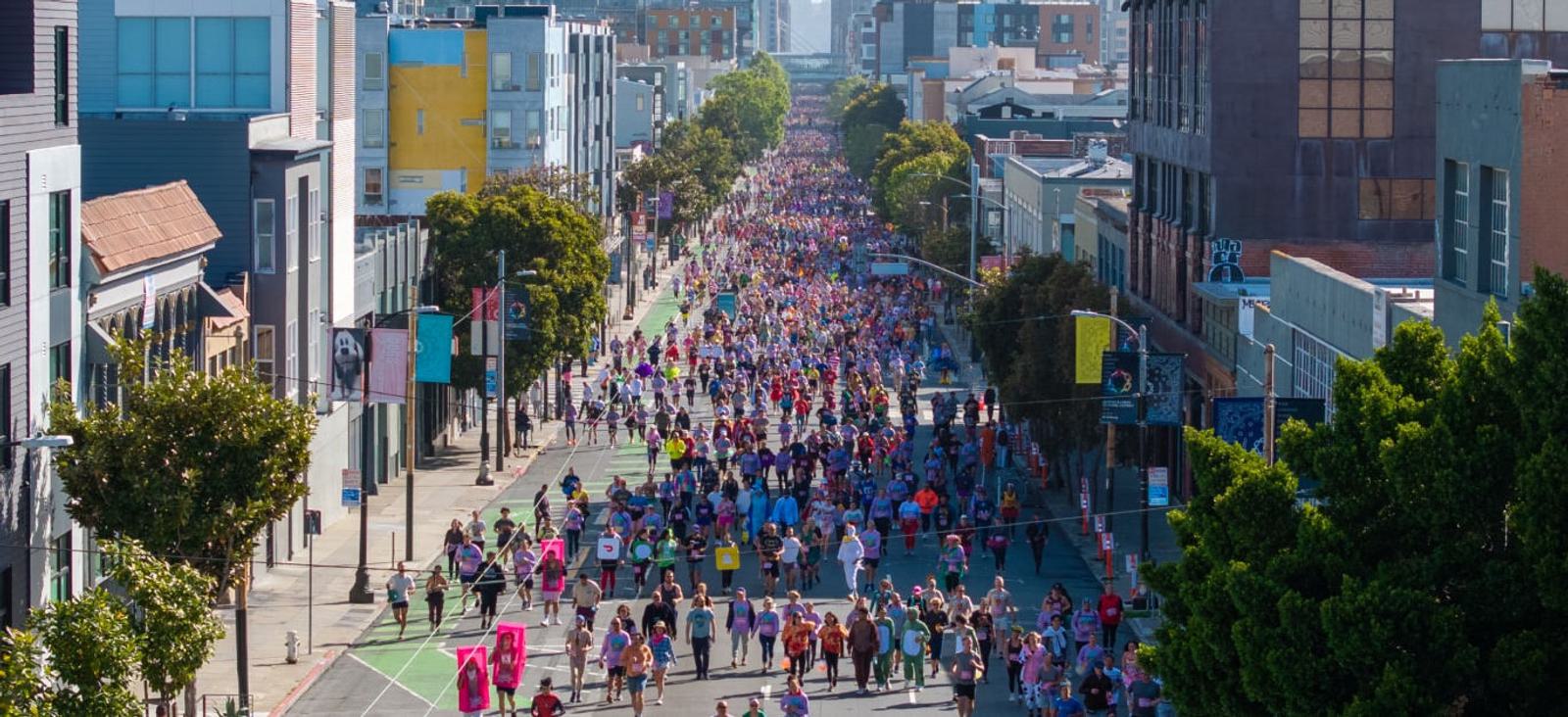 A large crowd of people participates in a street marathon or parade, filling a city street lined with buildings and trees on a sunny day.