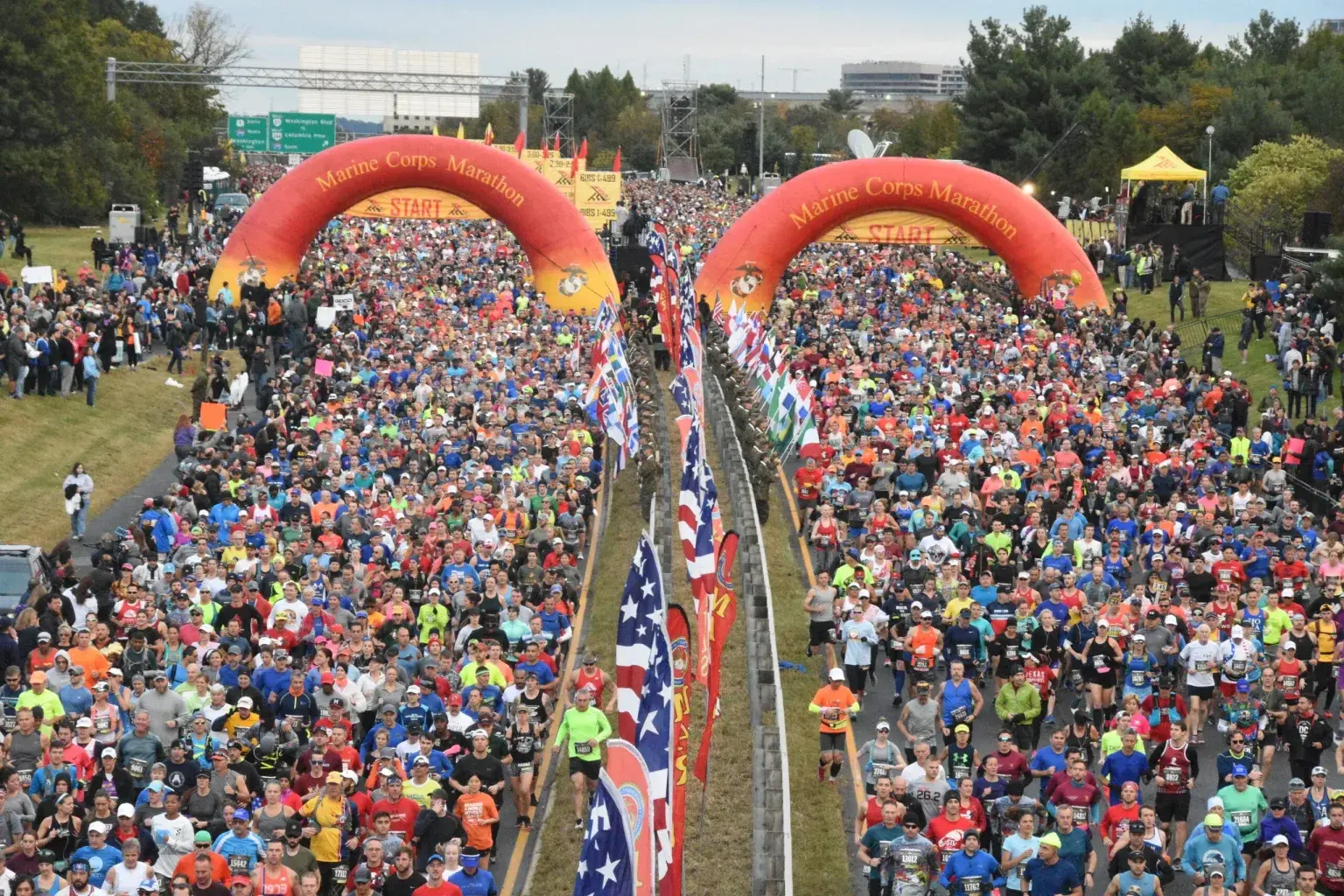 A large crowd of runners gathers at the starting line under two red inflatable arches labeled "Marine Corps Marathon," with rows of American flags lining the center and spectators watching from the sides.