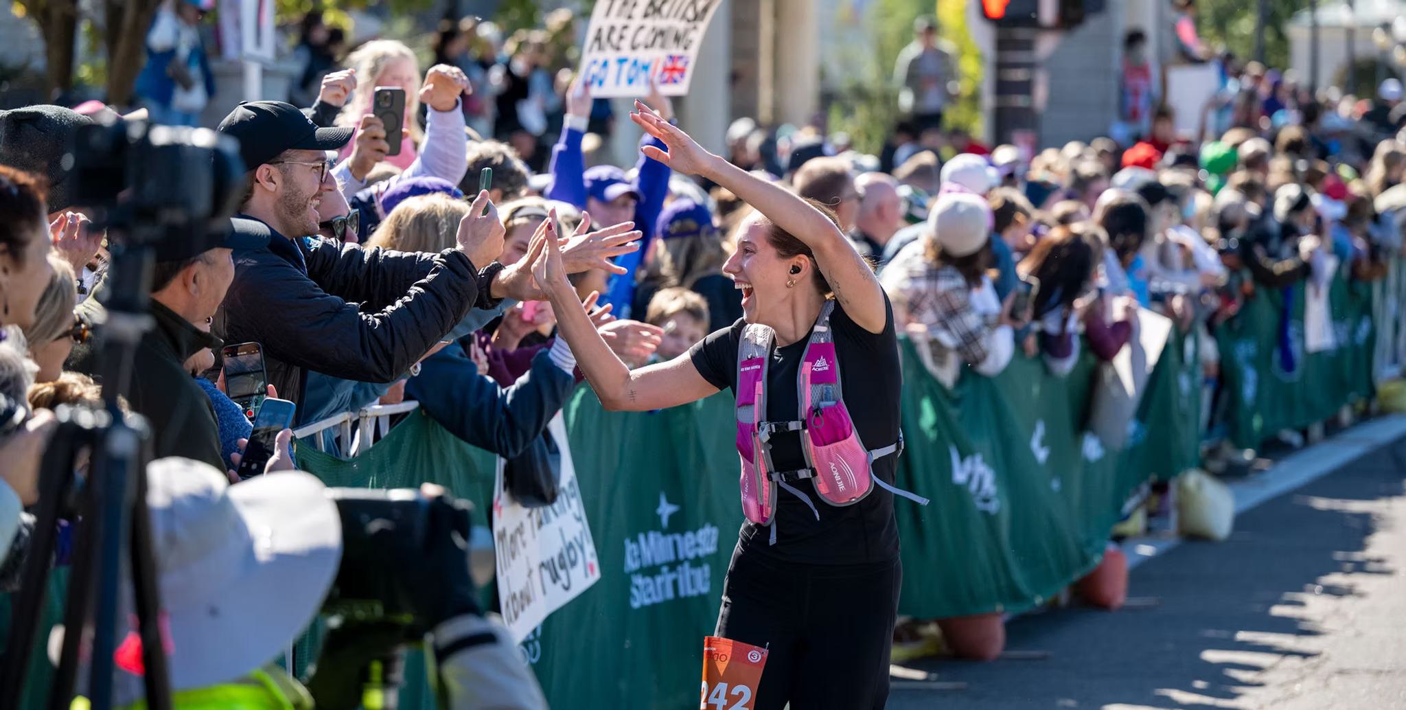 A marathon runner high-fives cheering spectators along a crowded racecourse, with people smiling, holding signs, and taking photos as she passes by in the sunlight.