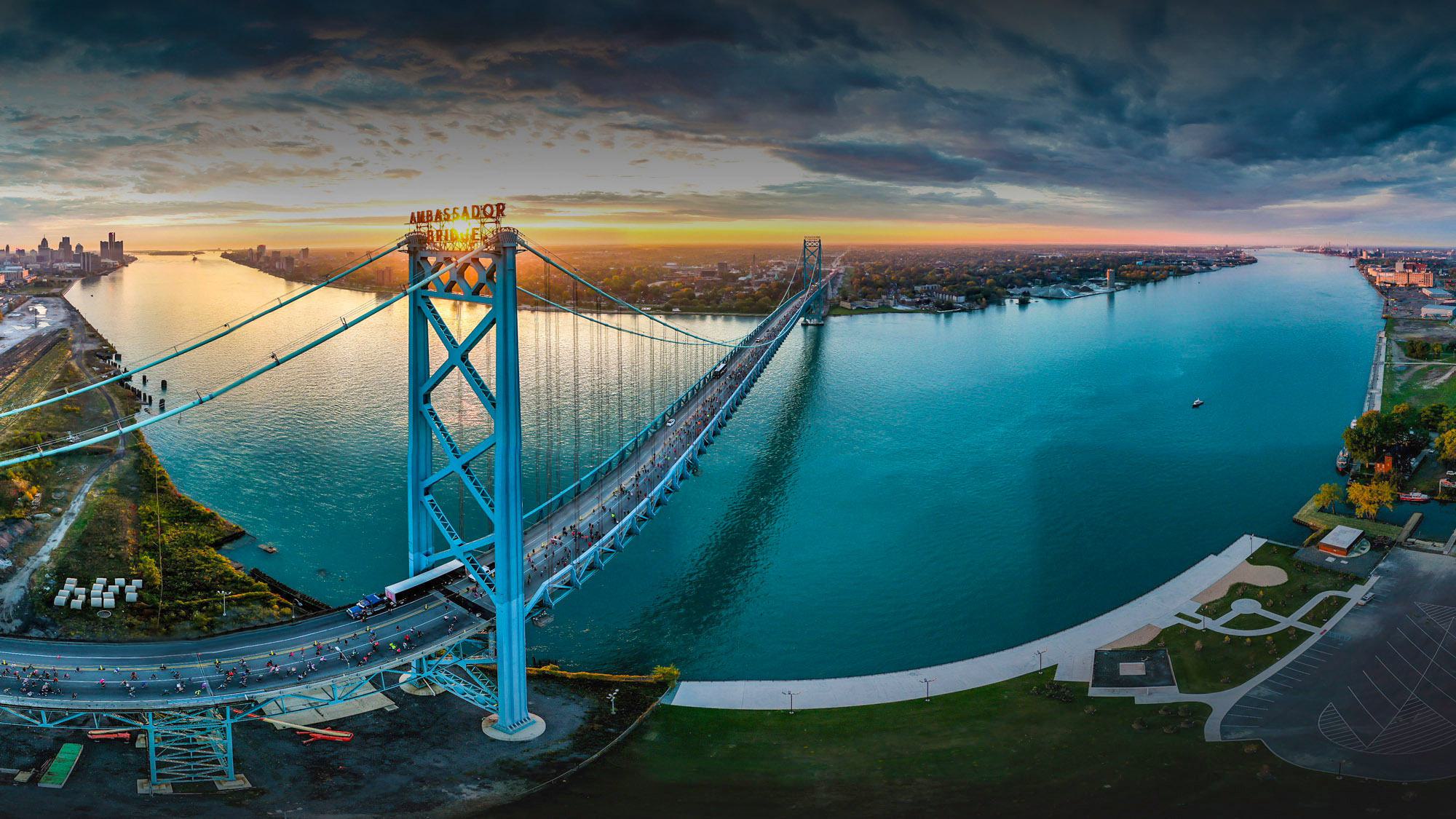 Aerial view of the Ambassador Bridge at sunset, spanning over the blue Detroit River, connecting Detroit, Michigan (left) and Windsor, Ontario (right), with city skylines visible in the distance.