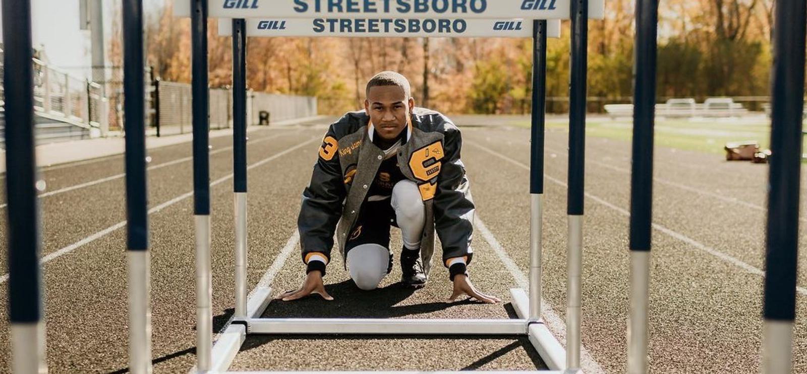 A person kneels at a track starting position under a hurdle with the word "Streetsboro" visible on it. The person is wearing a letterman jacket and appears focused, preparing to start running on the track outside. Trees and bleachers are in the background.