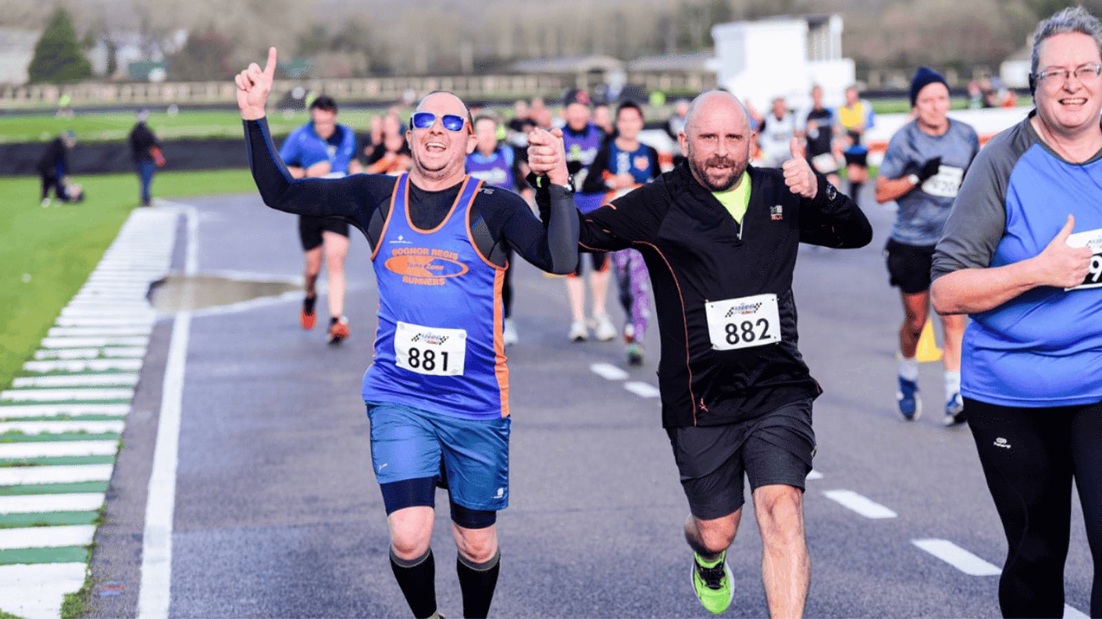 Two smiling men wearing race bibs numbered 881 and 882 hold hands and raise their arms in celebration as they run on a racetrack, surrounded by other runners in athletic gear.