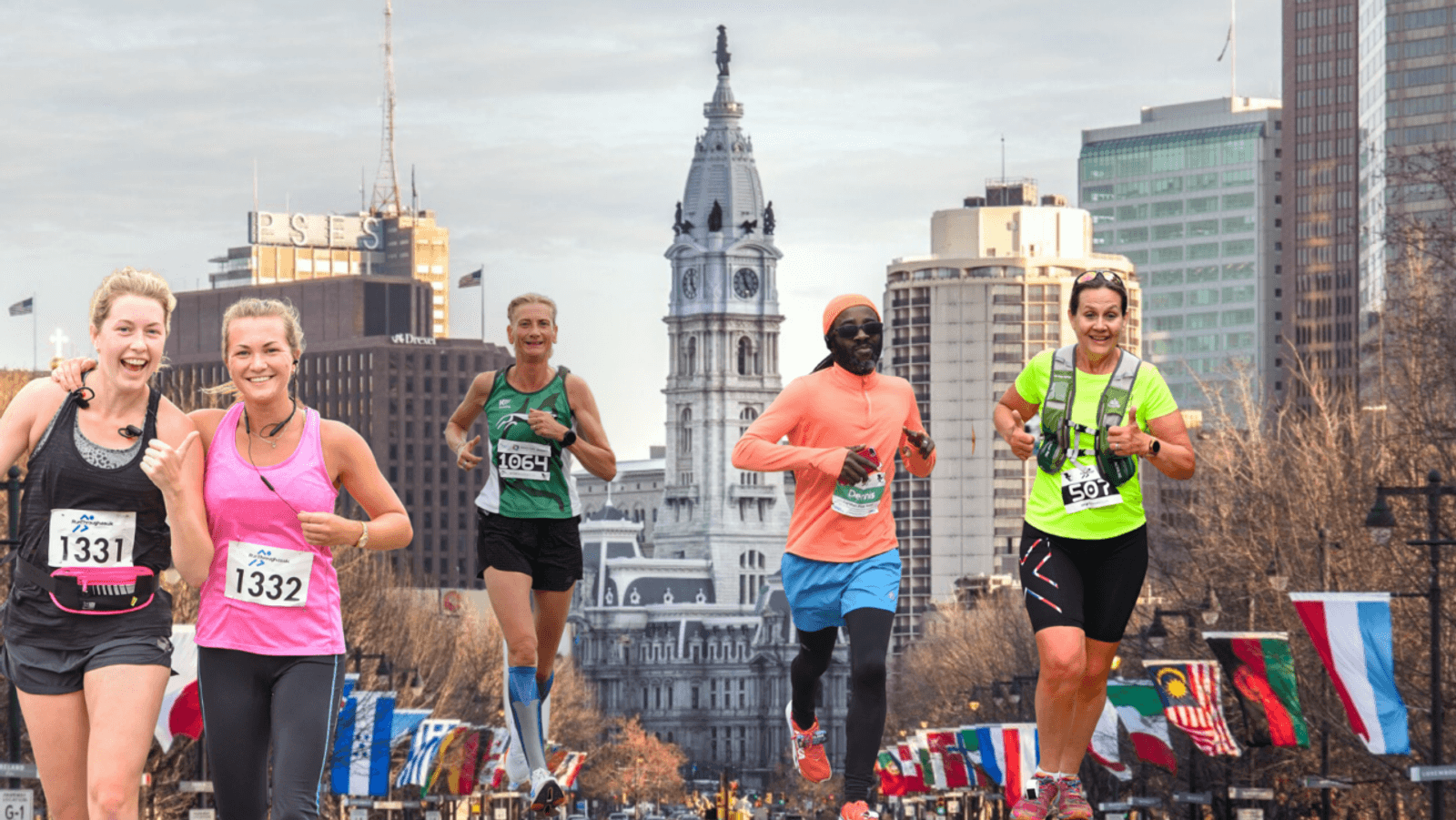 Five runners, smiling and wearing race bibs, run down a city street lined with flags and trees. A tall historic building with a clock tower rises in the background under a cloudy sky.