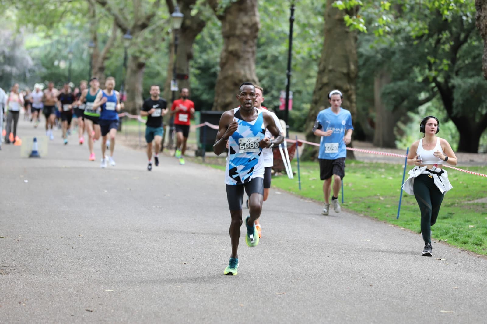 A group of runners participating in a race through a tree-lined park, with one runner in a blue and white outfit leading the group, and spectators and participants visible in the background.