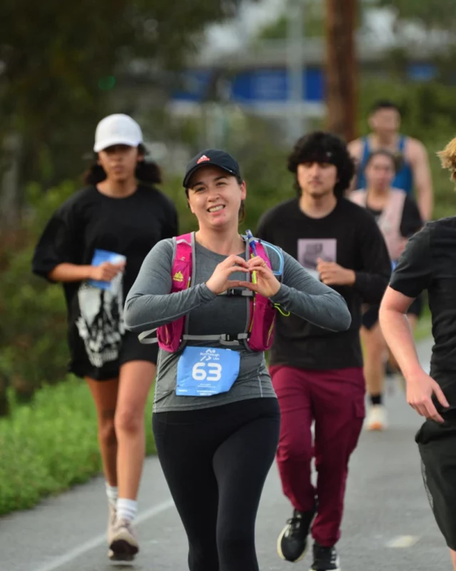 A woman wearing a race bib labeled 63 runs outdoors, smiling and making a heart shape with her hands. Several other runners are behind her on the path. It appears to be a race or organized run.