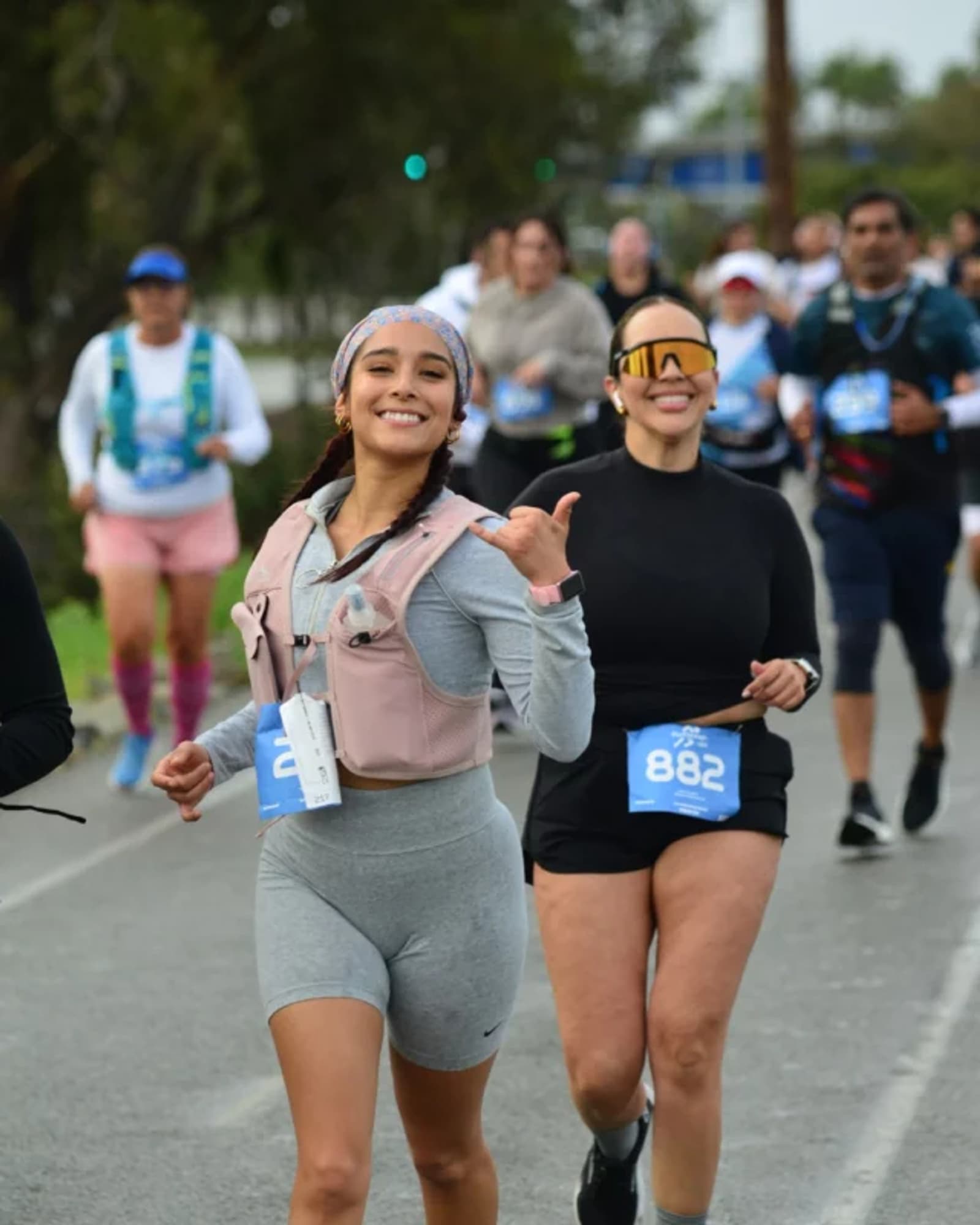 Two women in athletic wear, smiling and running in a race with numbered bibs among a group of participants on a road. The woman in front is making a shaka hand gesture and looking at the camera.