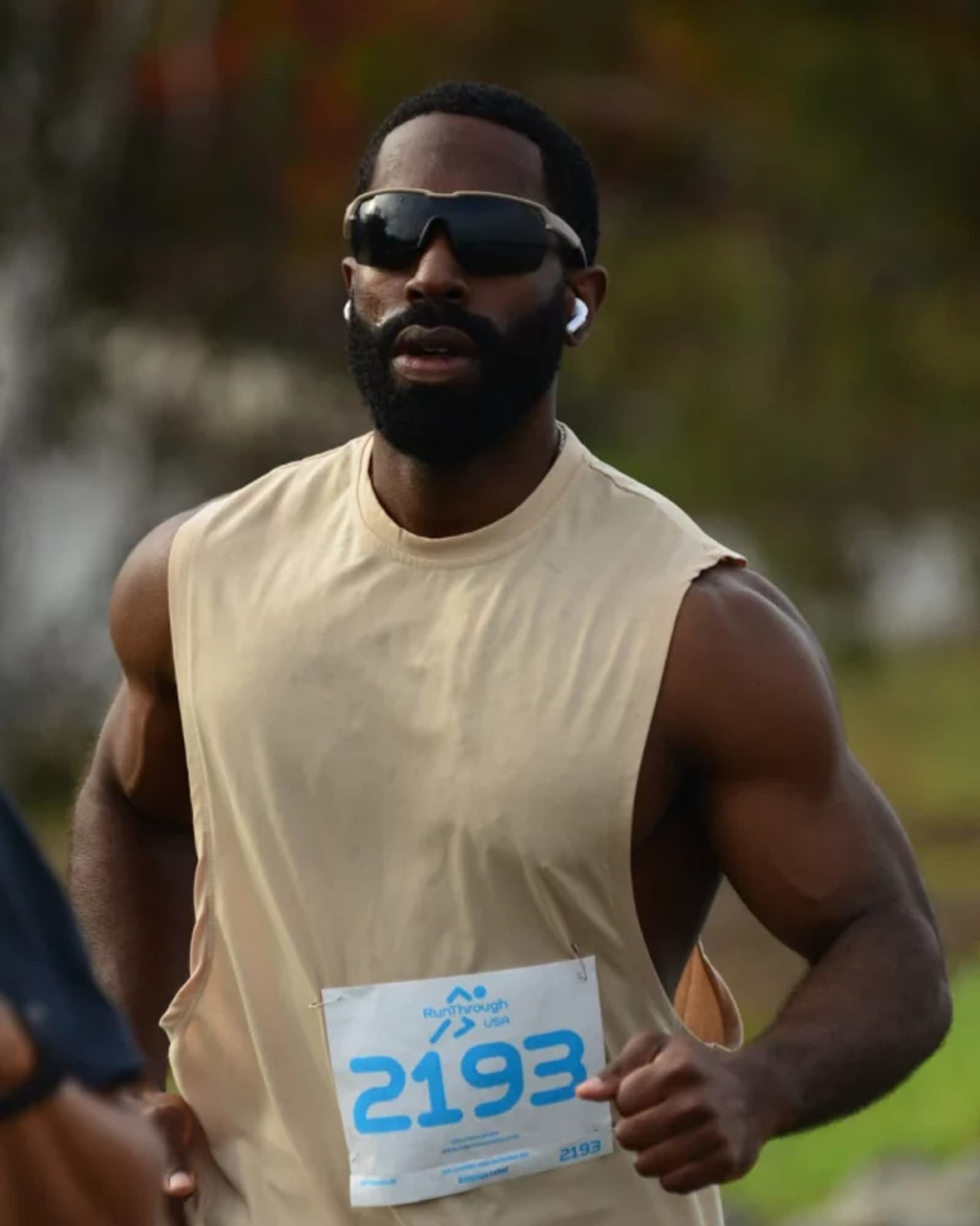 A man wearing sunglasses, wireless earbuds, and a sleeveless beige shirt runs outdoors with a race bib numbered 2193 pinned to his chest.