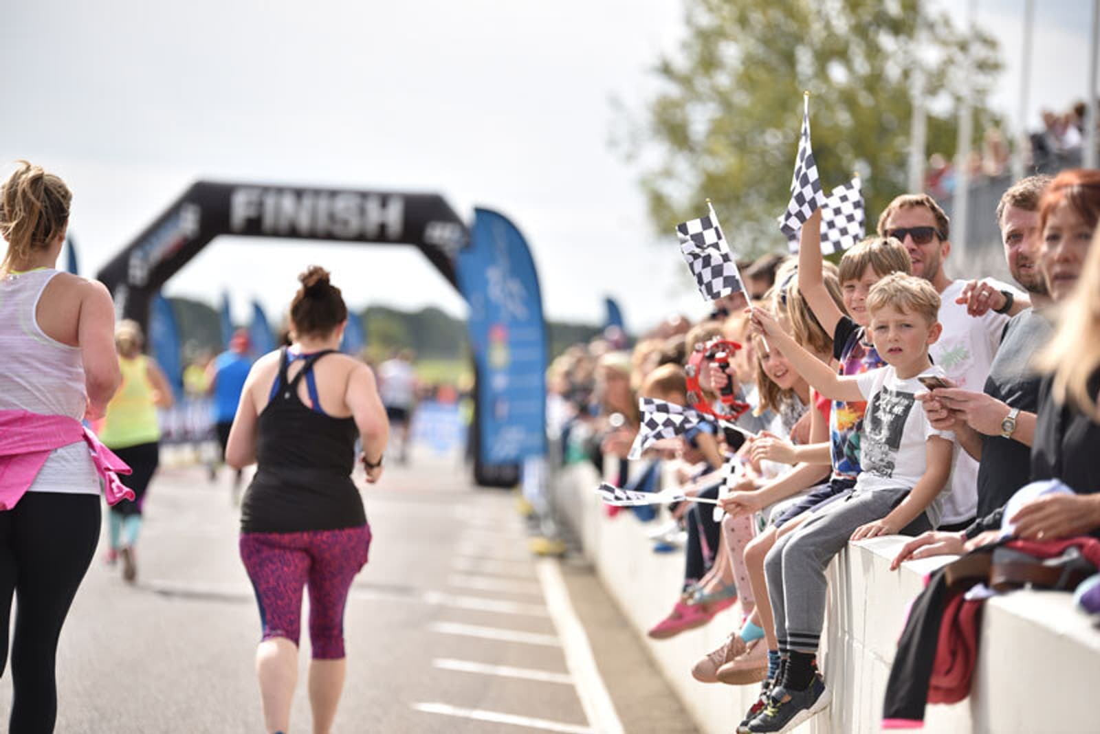 Runners approach the finish line as a crowd of spectators, including children waving checkered flags, cheer them on from the sidelines at a sunny outdoor event.