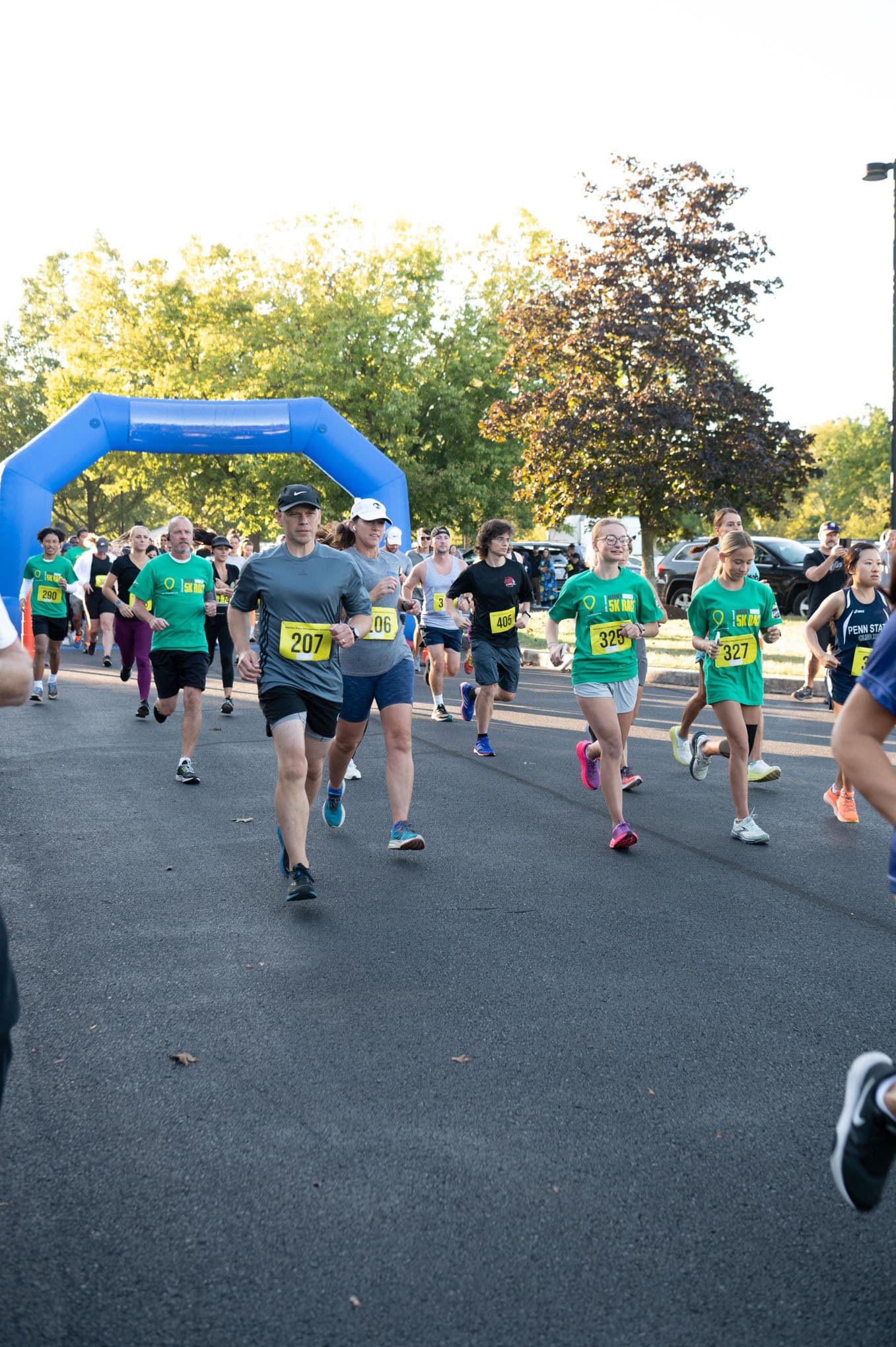 Runners wearing race bibs participate in an outdoor race, passing under a blue inflatable archway on a sunny day, with trees and spectators in the background.