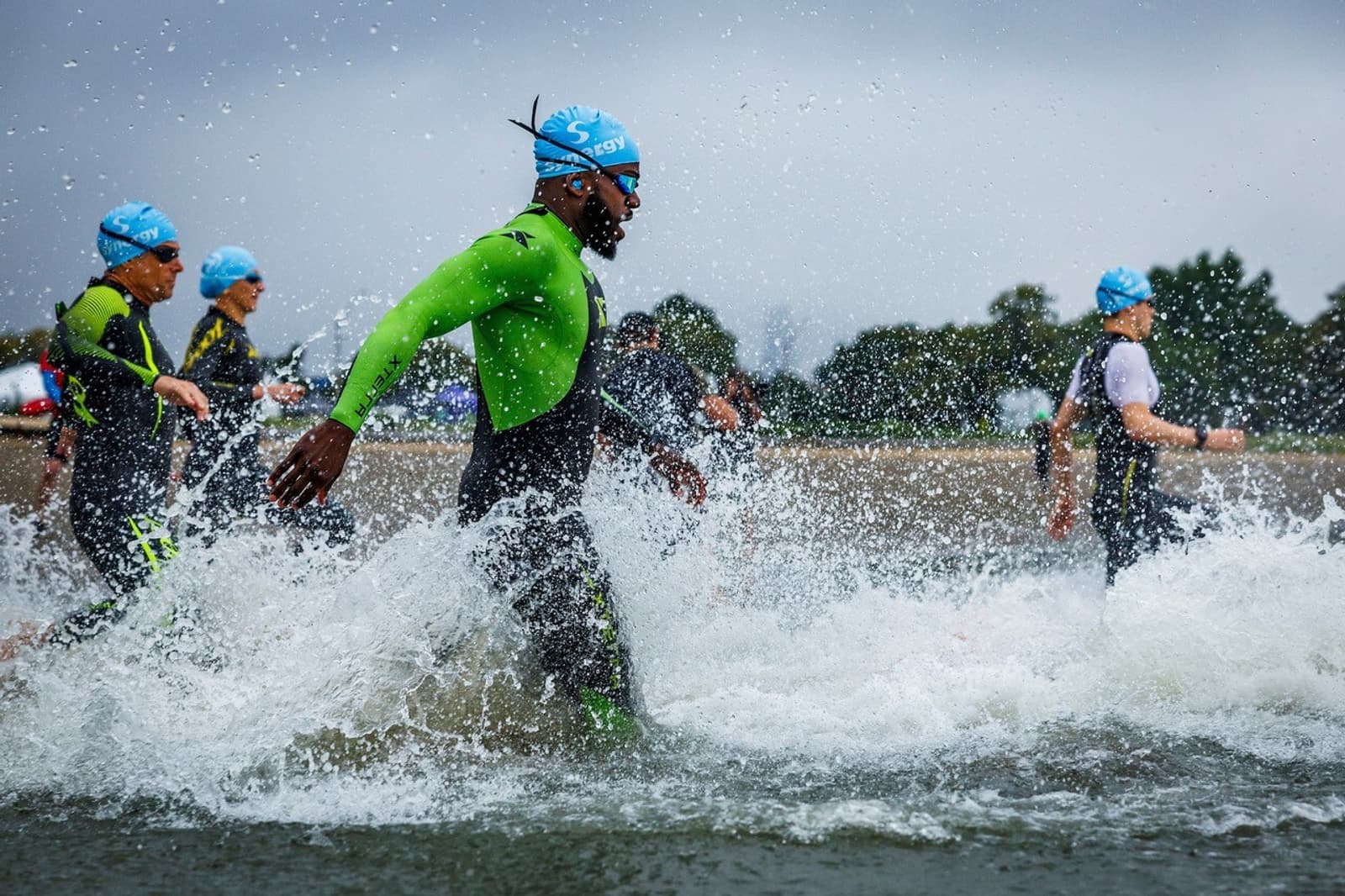 A group of athletes wearing wetsuits and blue swim caps run through shallow water at the start of a triathlon race, splashing as they move forward under a cloudy sky.