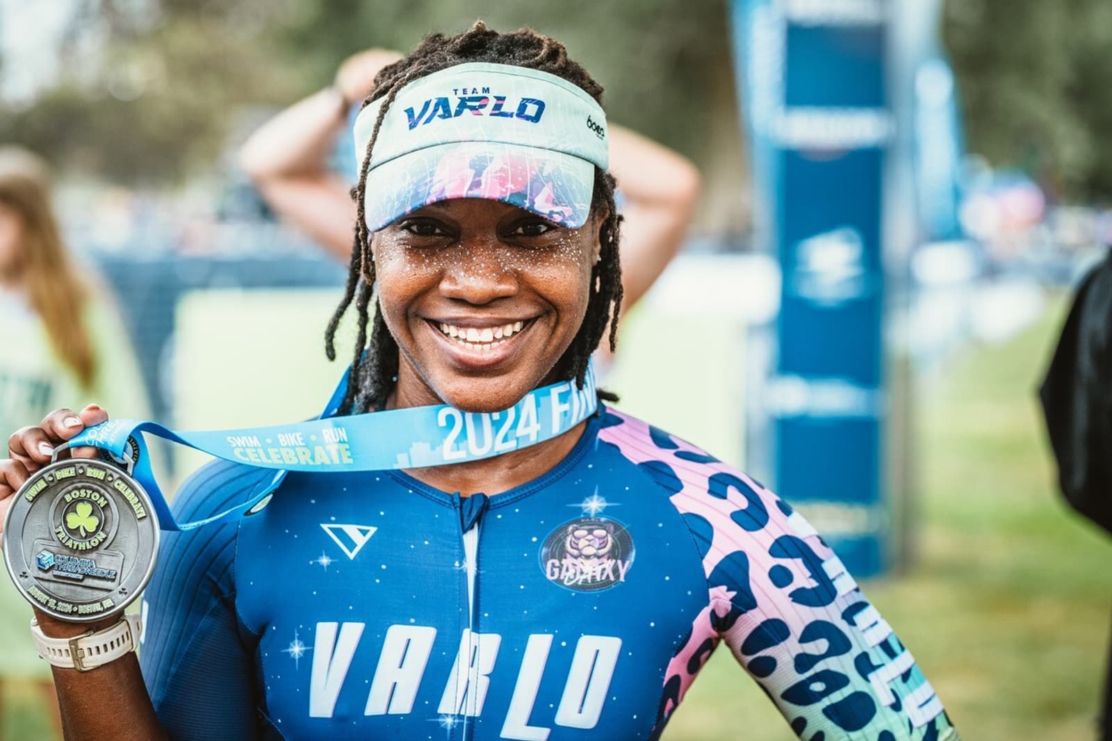A smiling athlete wearing a blue and pink VARLO suit and visor holds up a finisher’s medal after a race. She has braided hair and is standing outdoors with a blurred crowd and banners in the background.