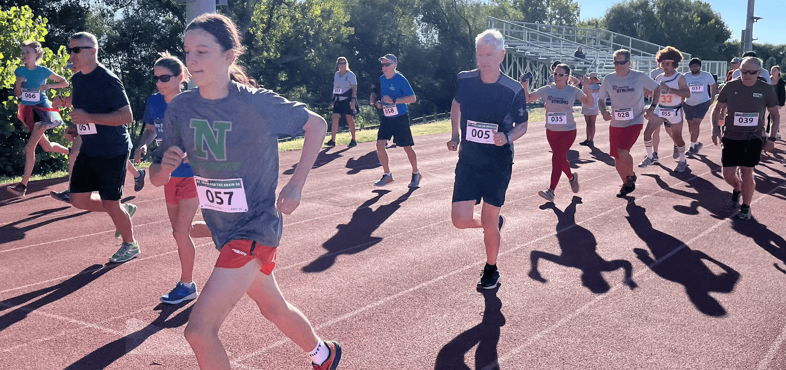 A group of runners wearing bib numbers start a race on an outdoor track under bright sunlight, casting long shadows on the red track surface. Trees and bleachers are visible in the background.