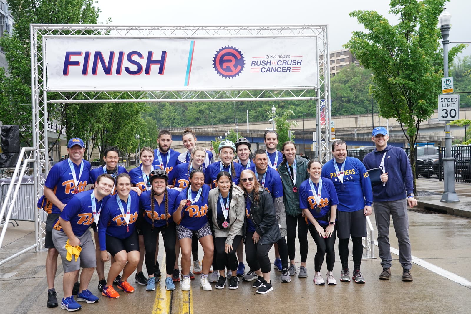 A group of people wearing matching blue "Pacers" shirts pose and smile together under a "FINISH" banner at an outdoor event, with medals around their necks and greenery in the background.