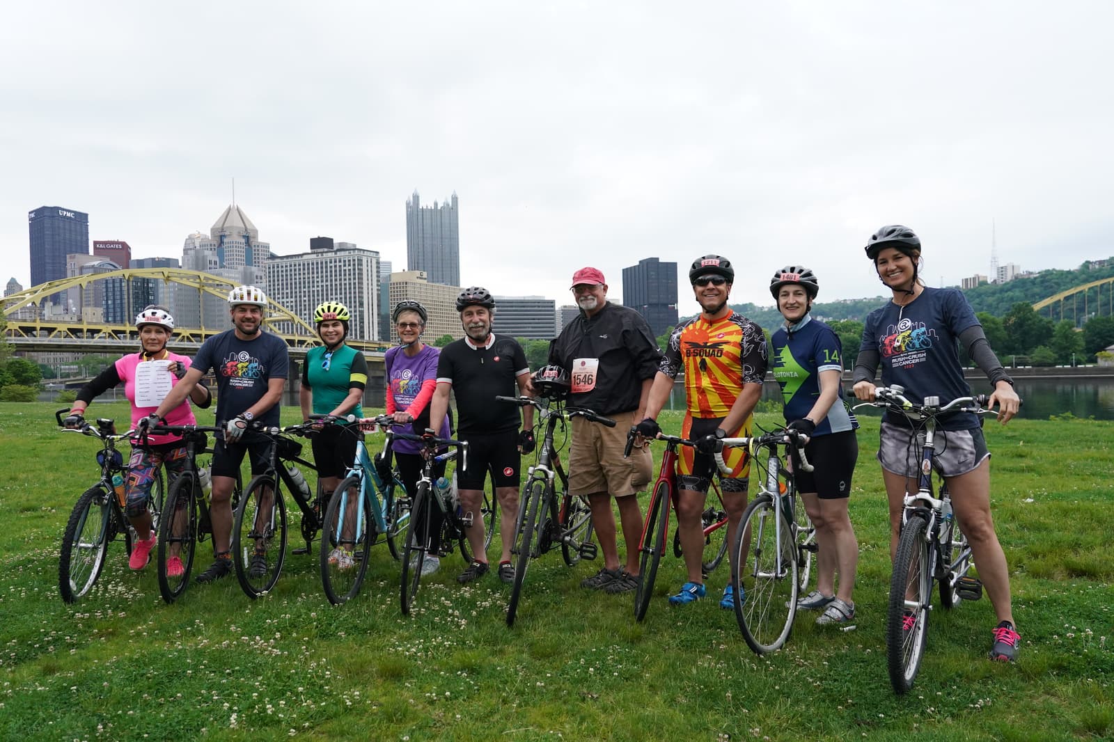 A group of nine cyclists and one man standing on grass with bikes, smiling at the camera. City buildings and yellow bridges are visible in the background on a cloudy day.