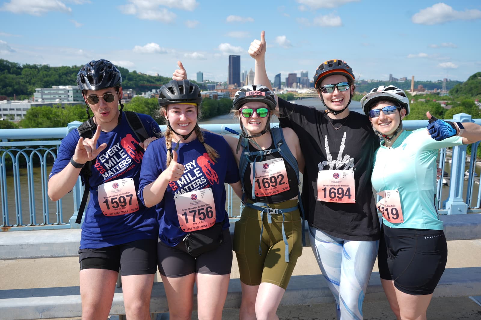 Five smiling cyclists wearing helmets and race bibs pose together on a bridge, giving thumbs up and peace signs, with a city skyline and green hills in the background on a sunny day.