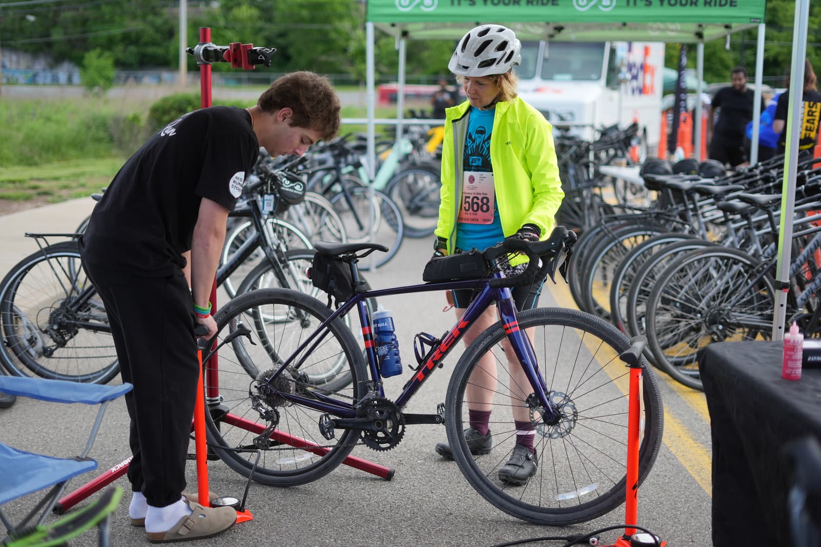 A person in a black shirt repairs a bicycle while another person in a bright yellow jacket and helmet watches at a bike event with many bicycles parked in the background.