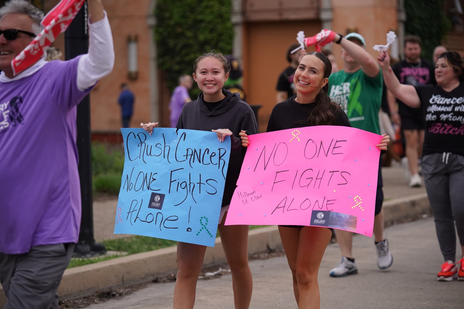 Two smiling people walk in a cancer awareness event holding signs that read "Crush Cancer, No One Fights Alone!" and "No One Fights Alone." Others walk and cheer behind them on a city street.