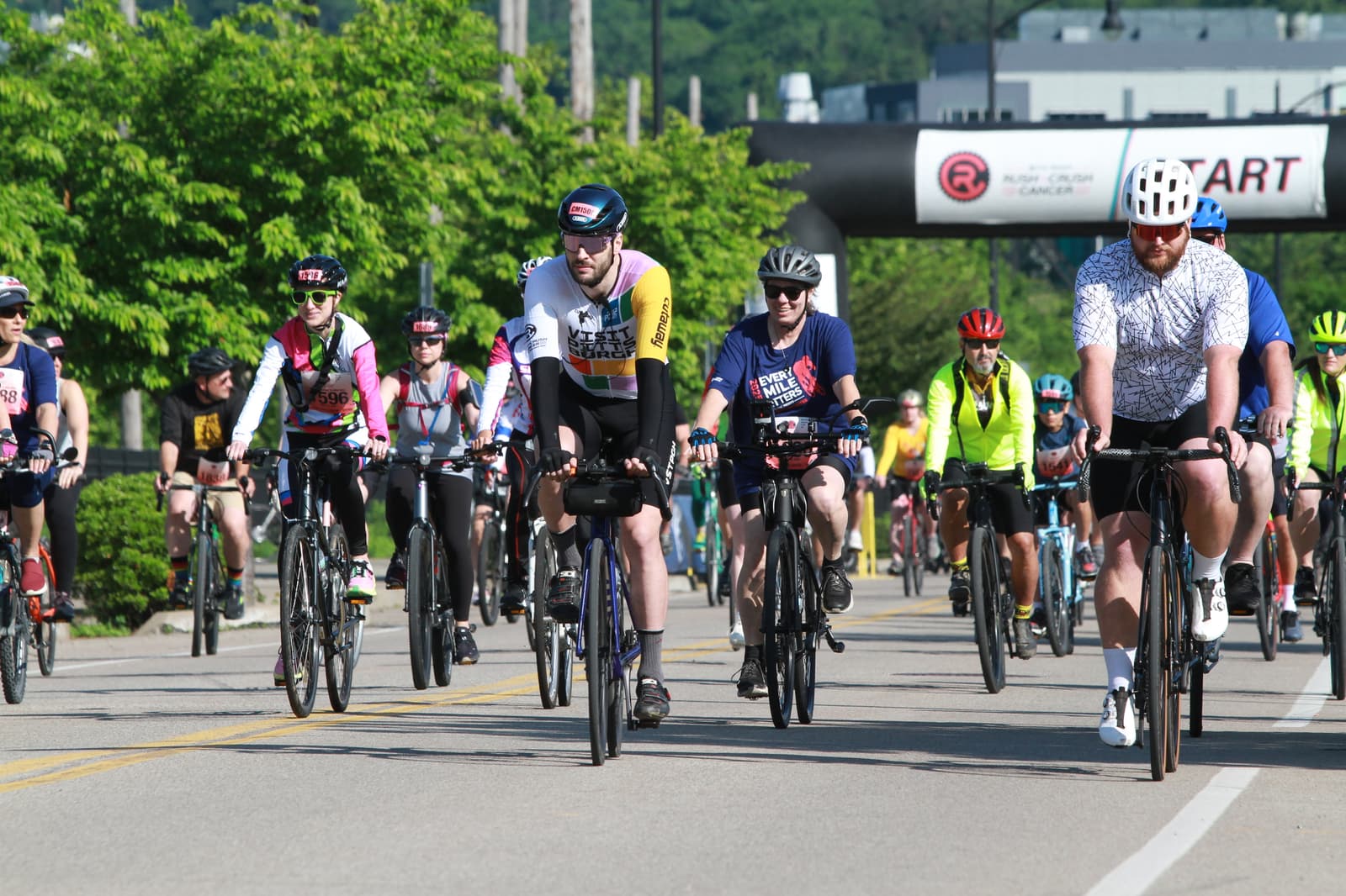 A group of cyclists wearing helmets and colorful jerseys ride together on a road during a cycling event, with a "START" banner visible in the background. Trees and buildings line the route.