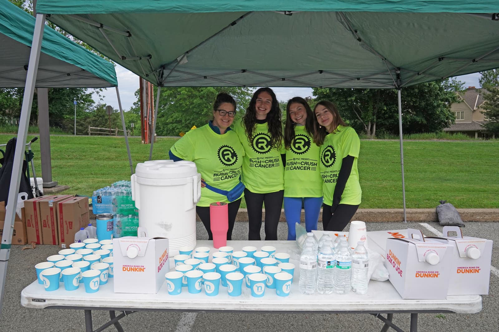 Four smiling people wearing matching neon green shirts stand behind a table with cups of water, water bottles, and boxes under a canopy tent at an outdoor event. Trees and grass are visible in the background.