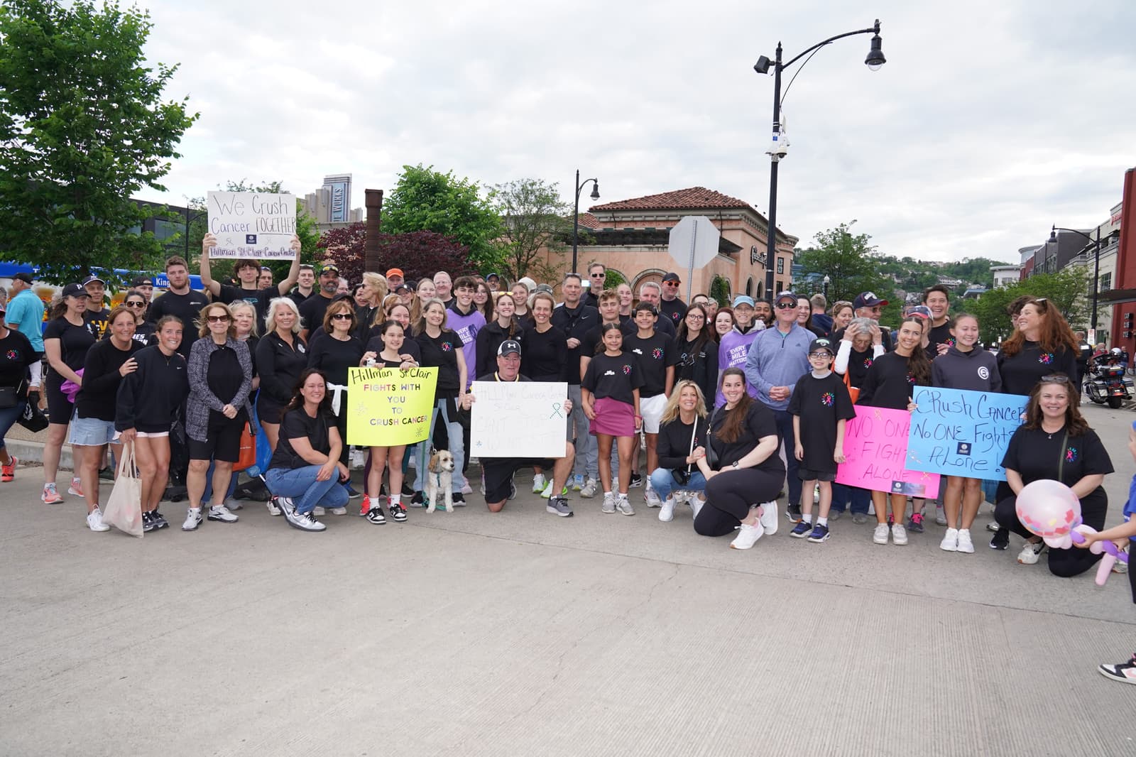 A large group of people pose together outdoors on a city street, many wearing matching shirts. Several hold colorful signs supporting cancer awareness and fundraising. Trees and buildings are visible in the background.