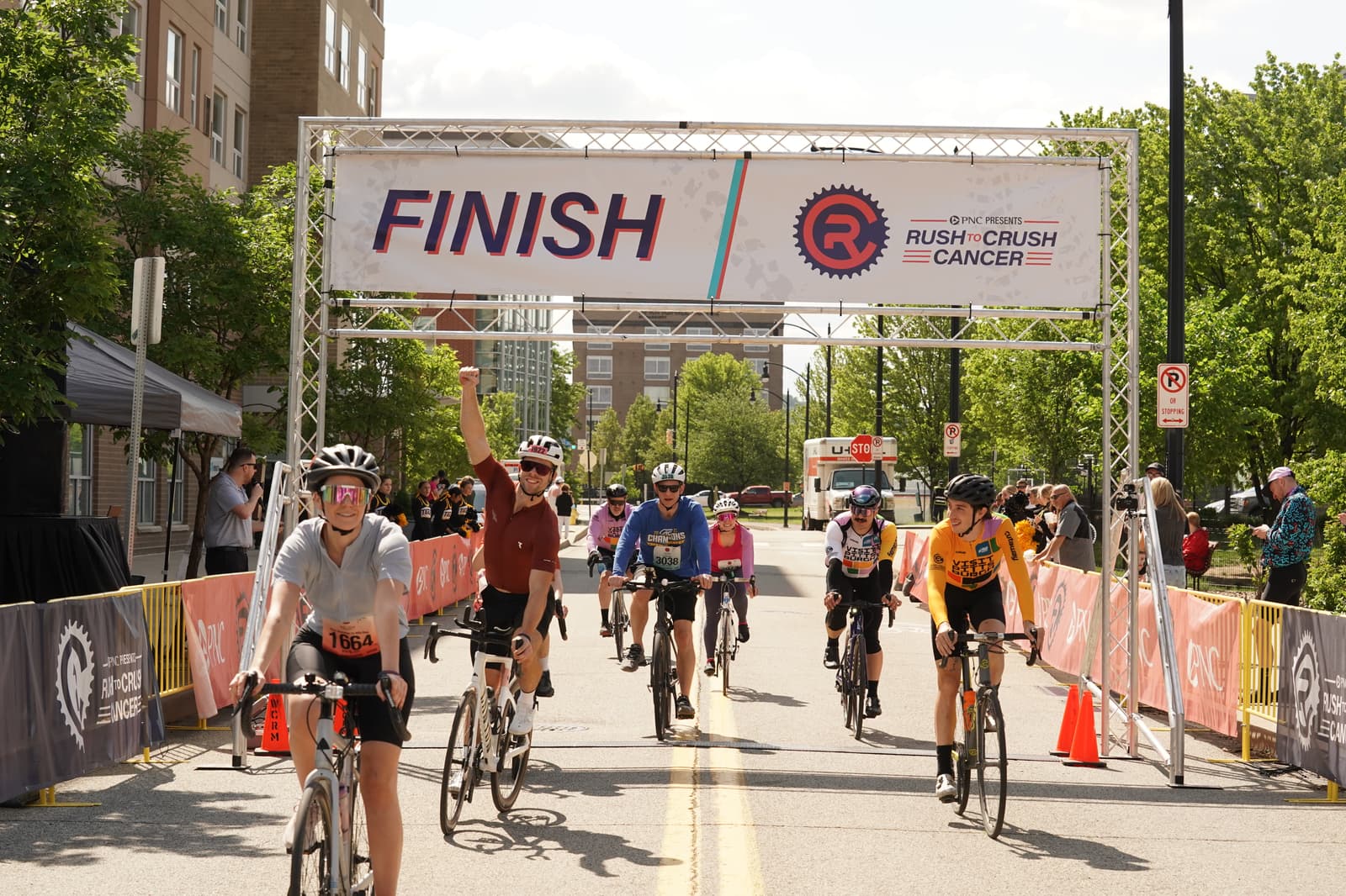 Cyclists cross the finish line at a road race event, with one rider raising an arm in celebration. A banner reads "FINISH" and "Rush Crush Cancer." Spectators and trees line the street on a sunny day.